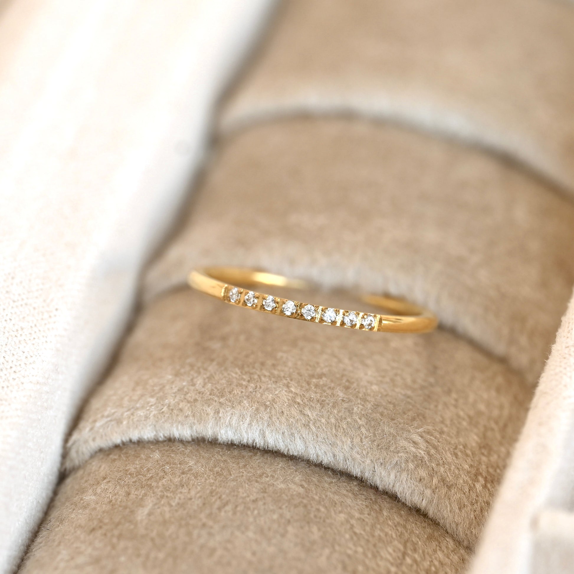 A delicate yellow gold band ring with a row of small clear gemstones, displayed on a beige velvet ring holder.