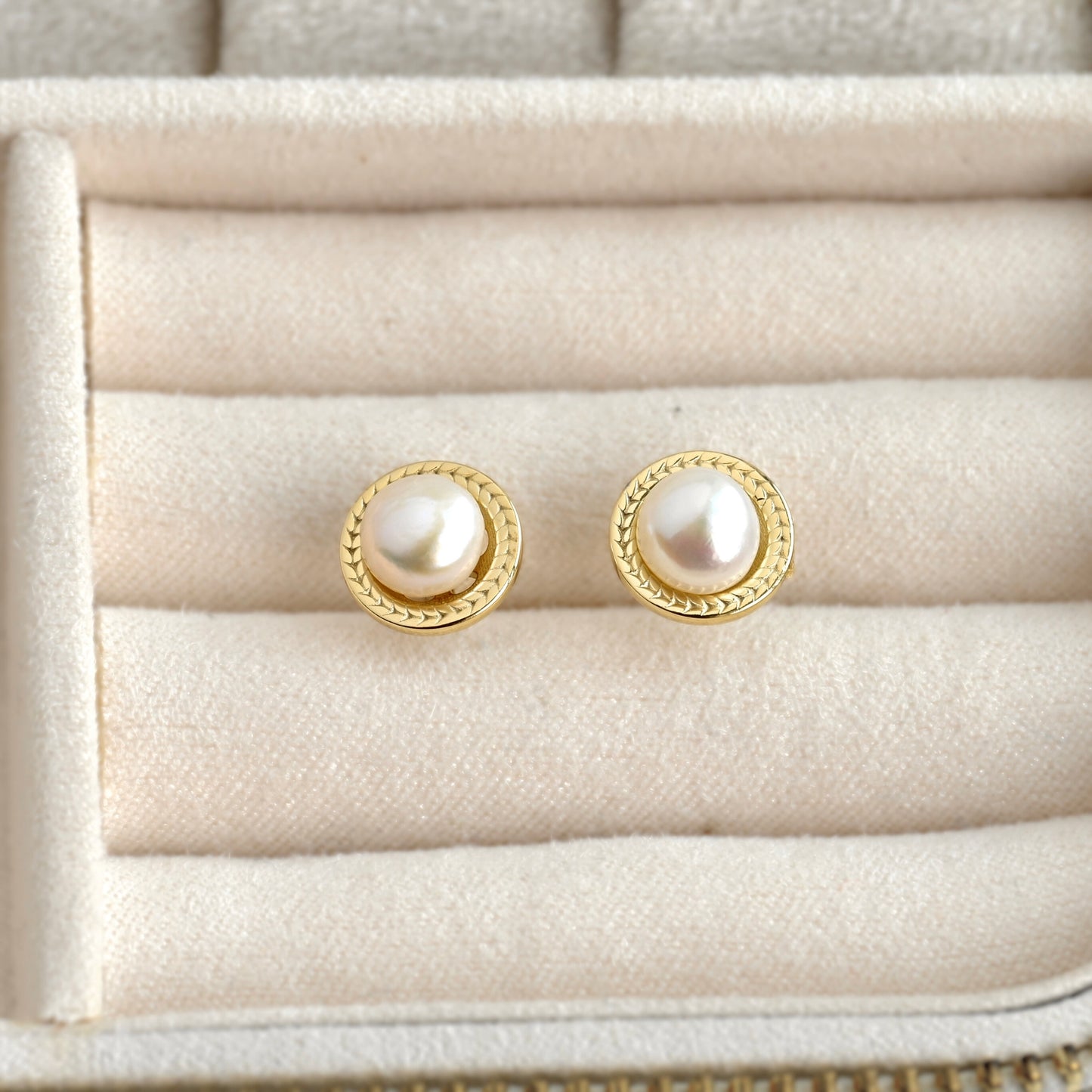 A pair of gold stud earrings with round white pearls, displayed on a cream-colored jewelry box.