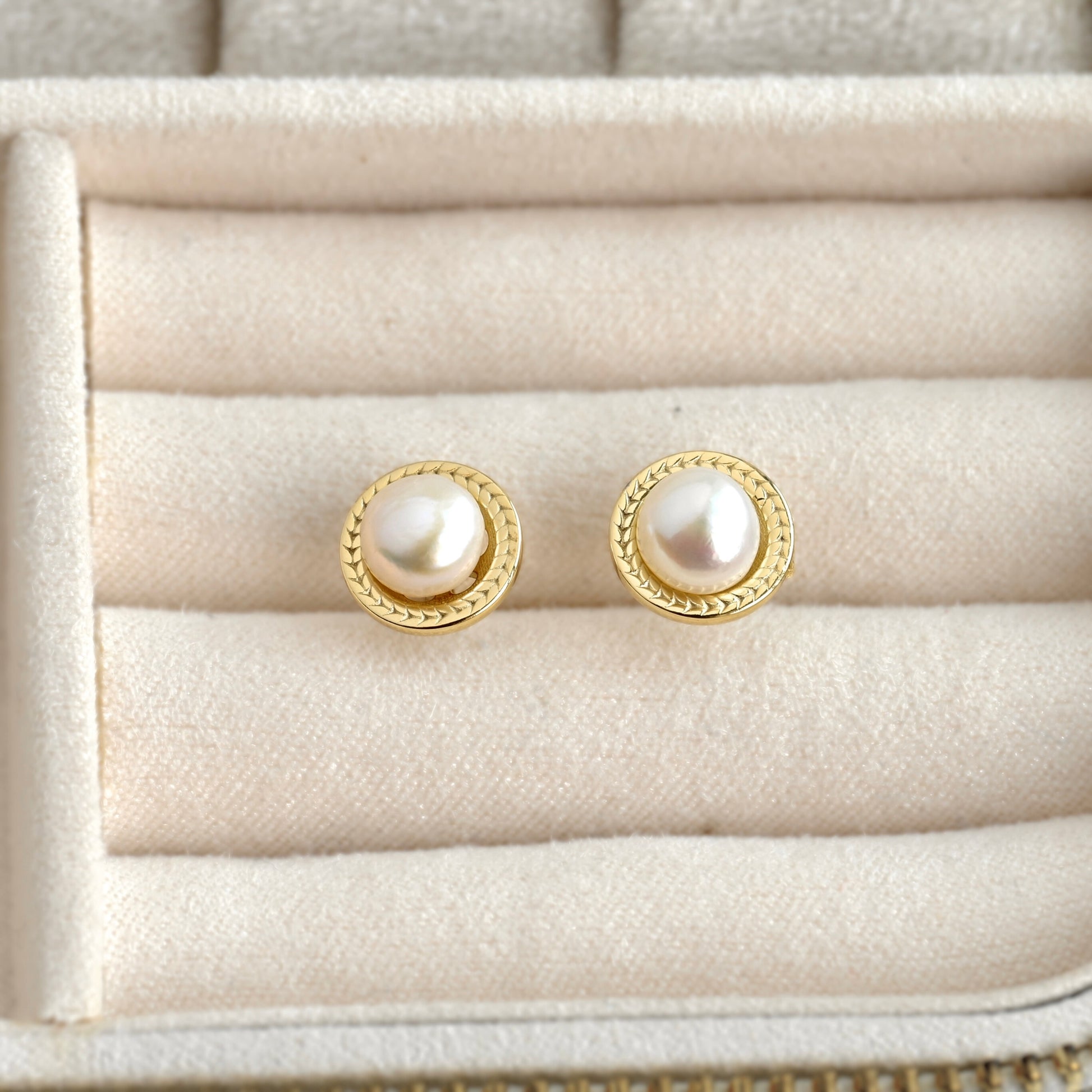 A pair of gold stud earrings with round white pearls, displayed on a cream-colored jewelry box.