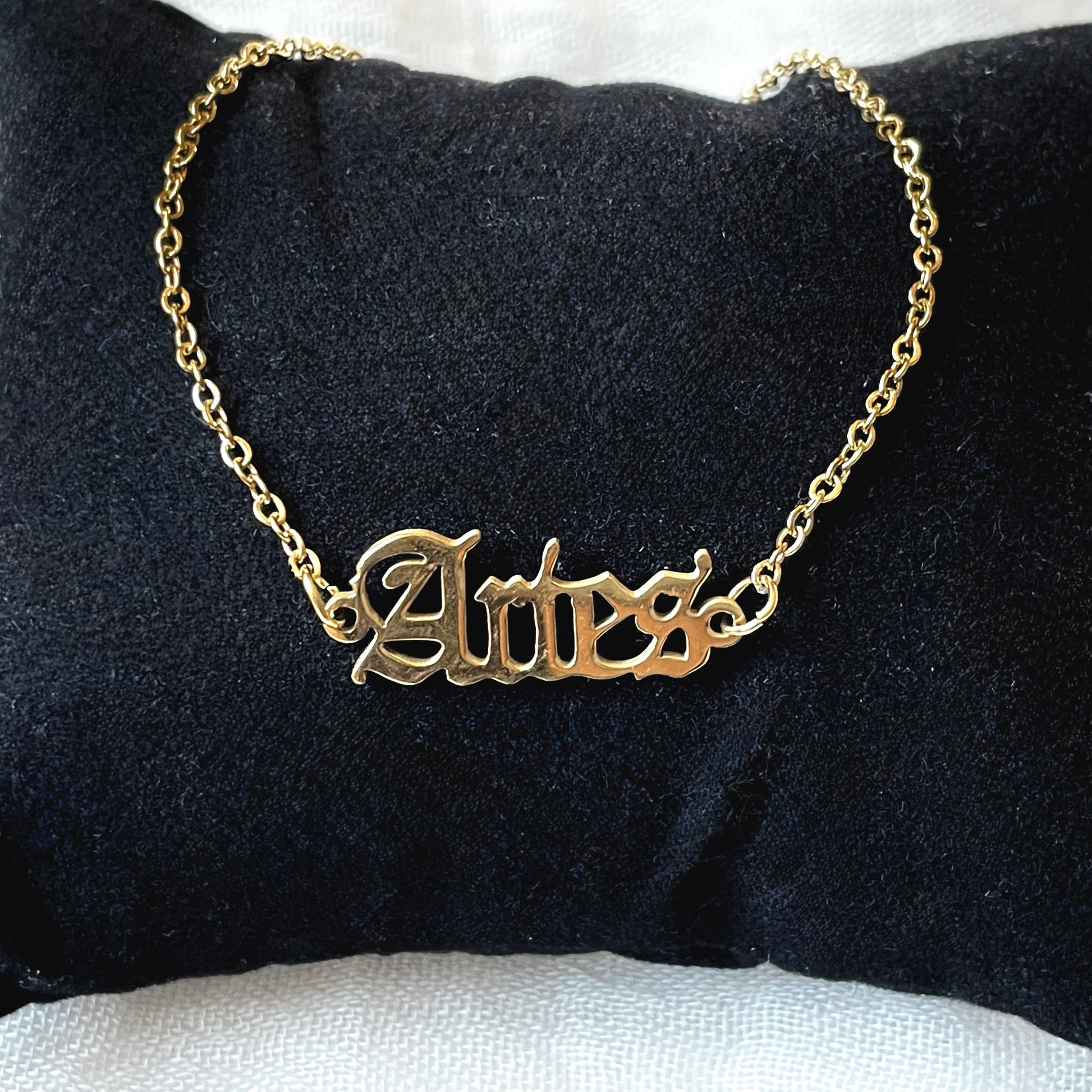 Gold zodiac ankle bracelet with 'Aries' in old english font on a black cushion