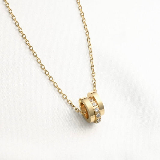 A gold chain necklace with a pendant consisting of three gold rings, one of which is set with small clear stones, displayed on a white fabric background.