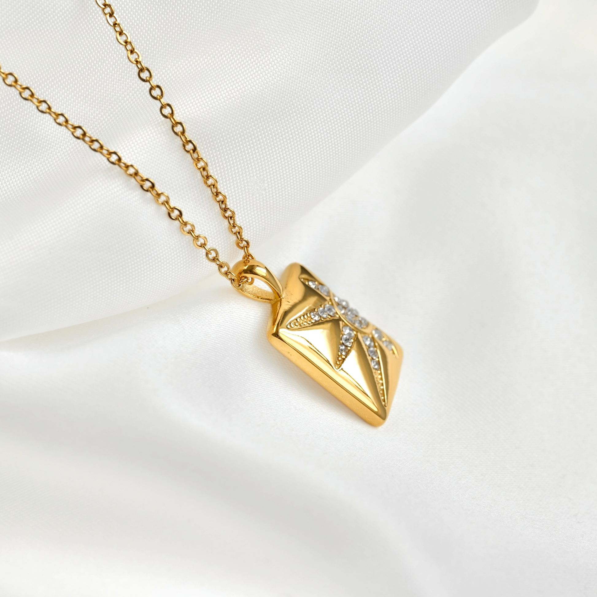 Side view of a gold pendant necklace featuring a geometric, starburst-like rectangular charm embellished with small clear stones, displayed on a soft white satin fabric background.