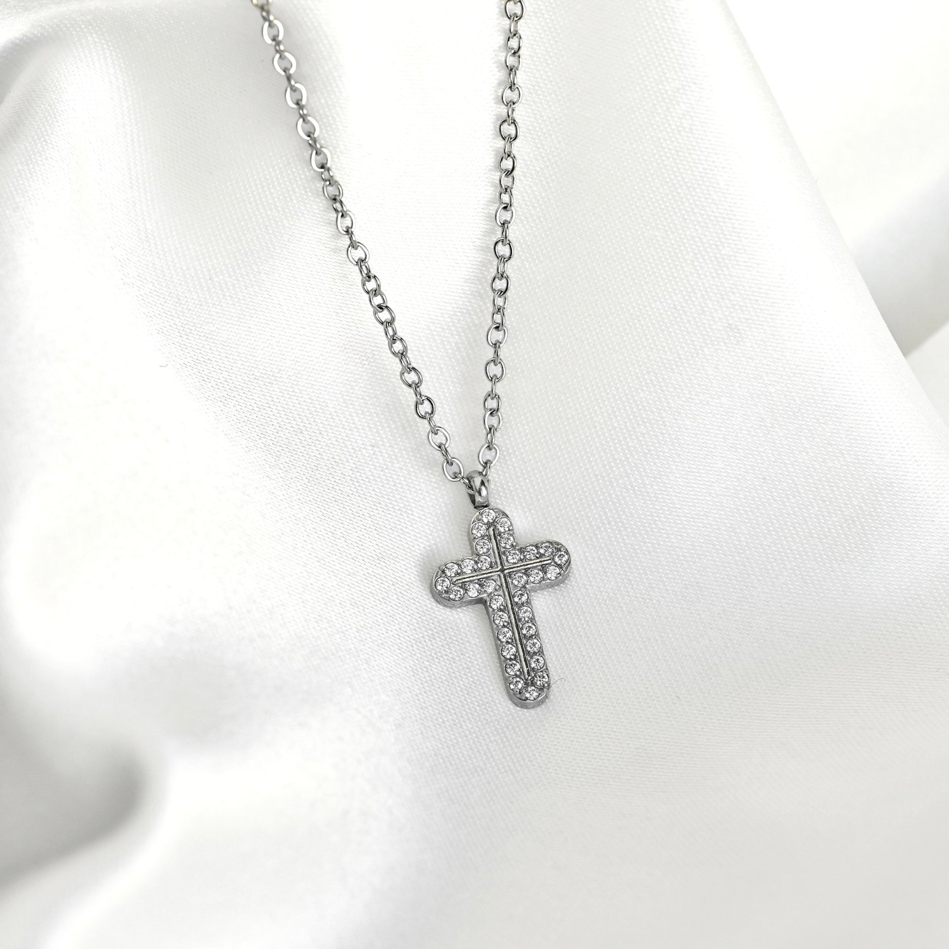 A silver cross pendant necklace paved with small clear stones, displayed on a draped white satin fabric background.