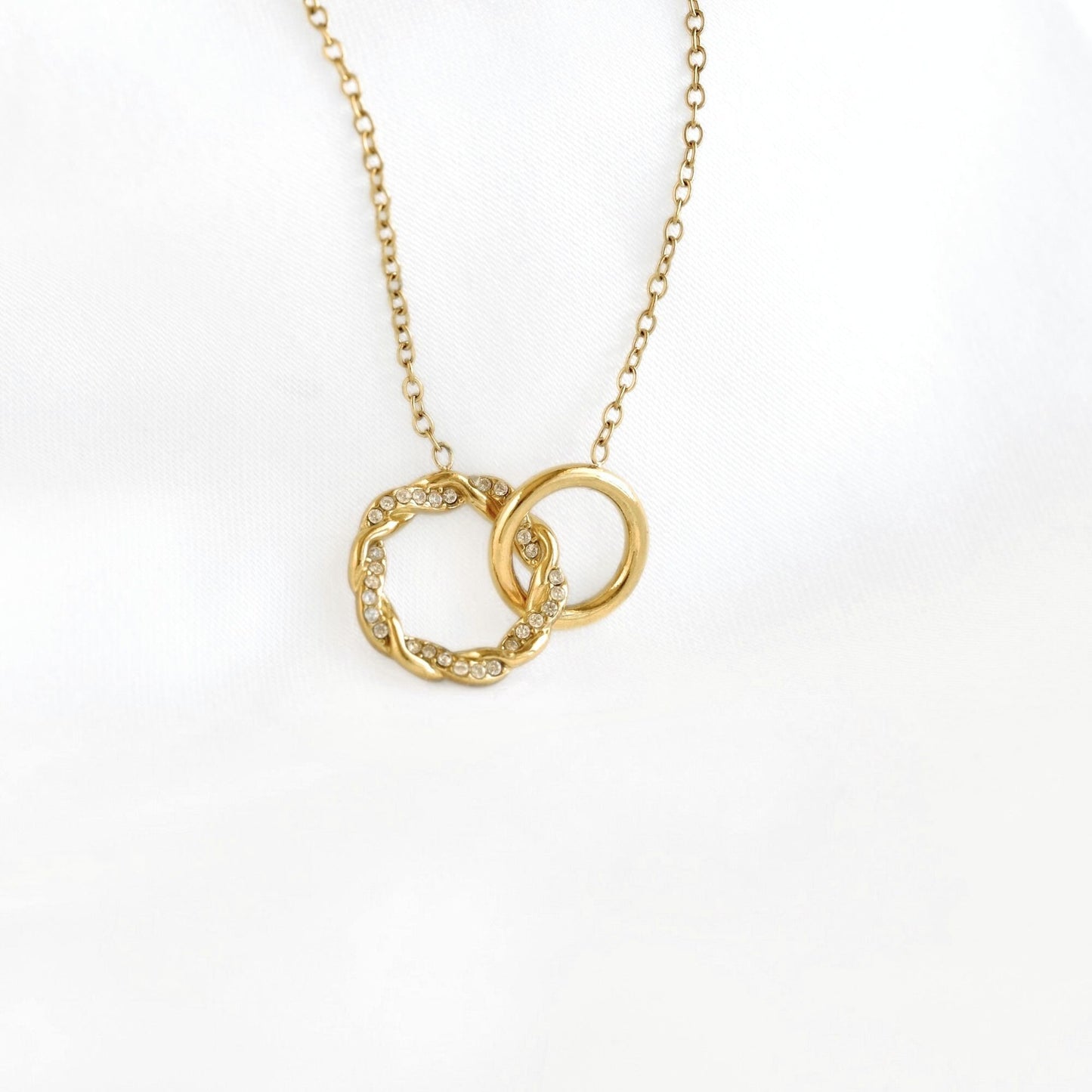 A gold chain necklace with an interlocking double-ring pendant, featuring one smooth ring and one textured ring set with small clear stones, displayed on a white fabric background.