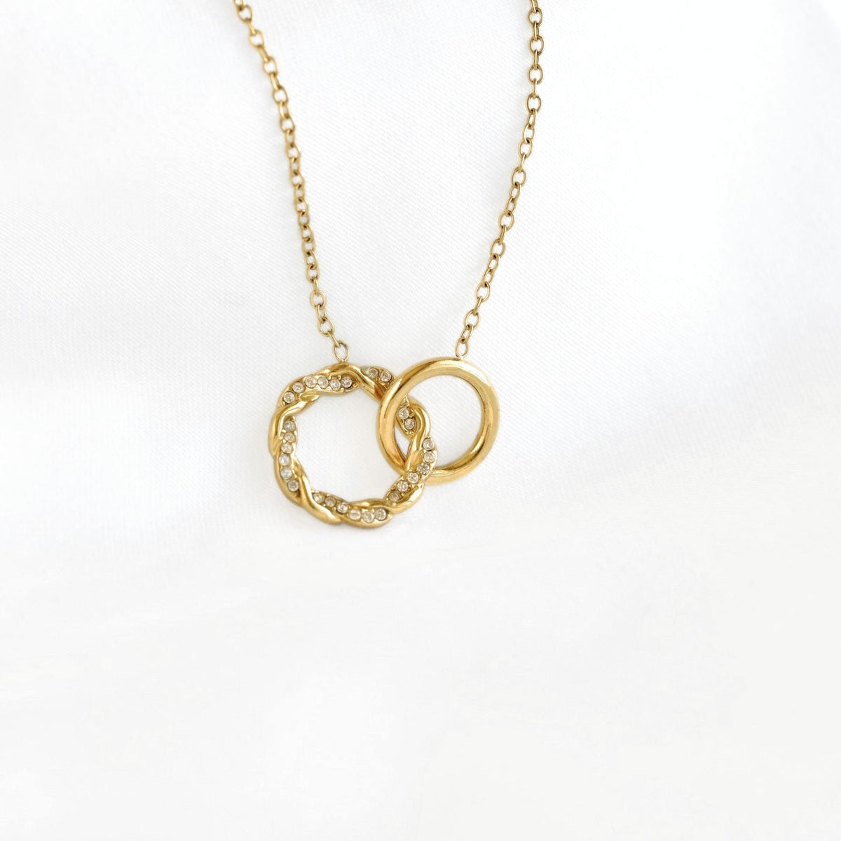 A gold chain necklace with an interlocking double-ring pendant, featuring one smooth ring and one textured ring set with small clear stones, displayed on a white fabric background.