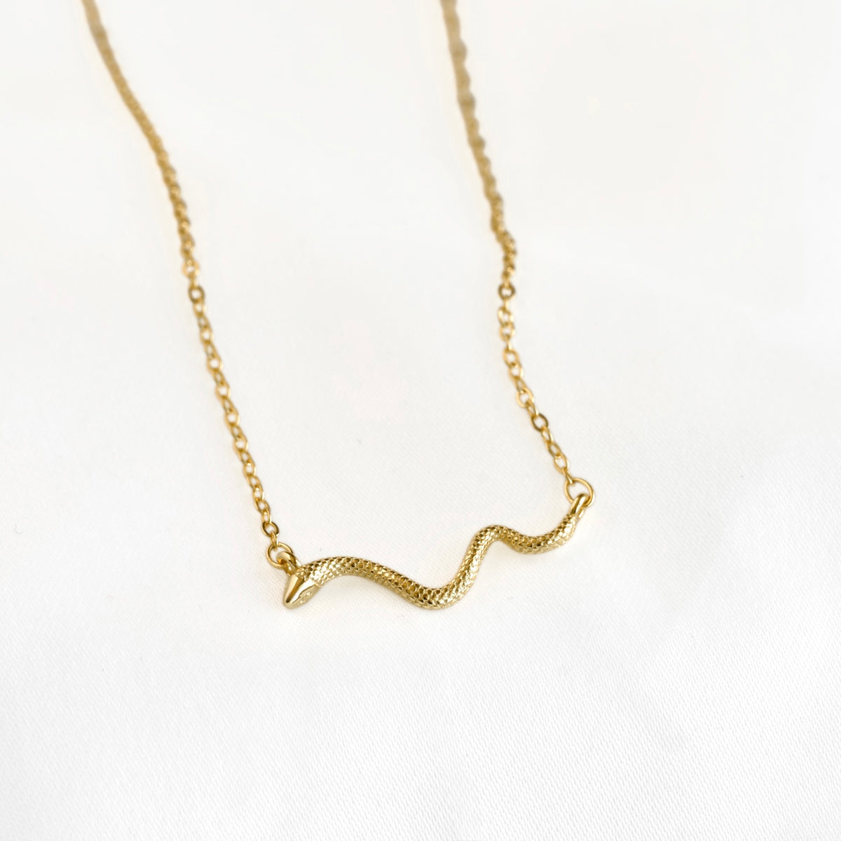 A delicate gold chain necklace featuring a small, textured snake pendant, displayed on a clean white fabric background.