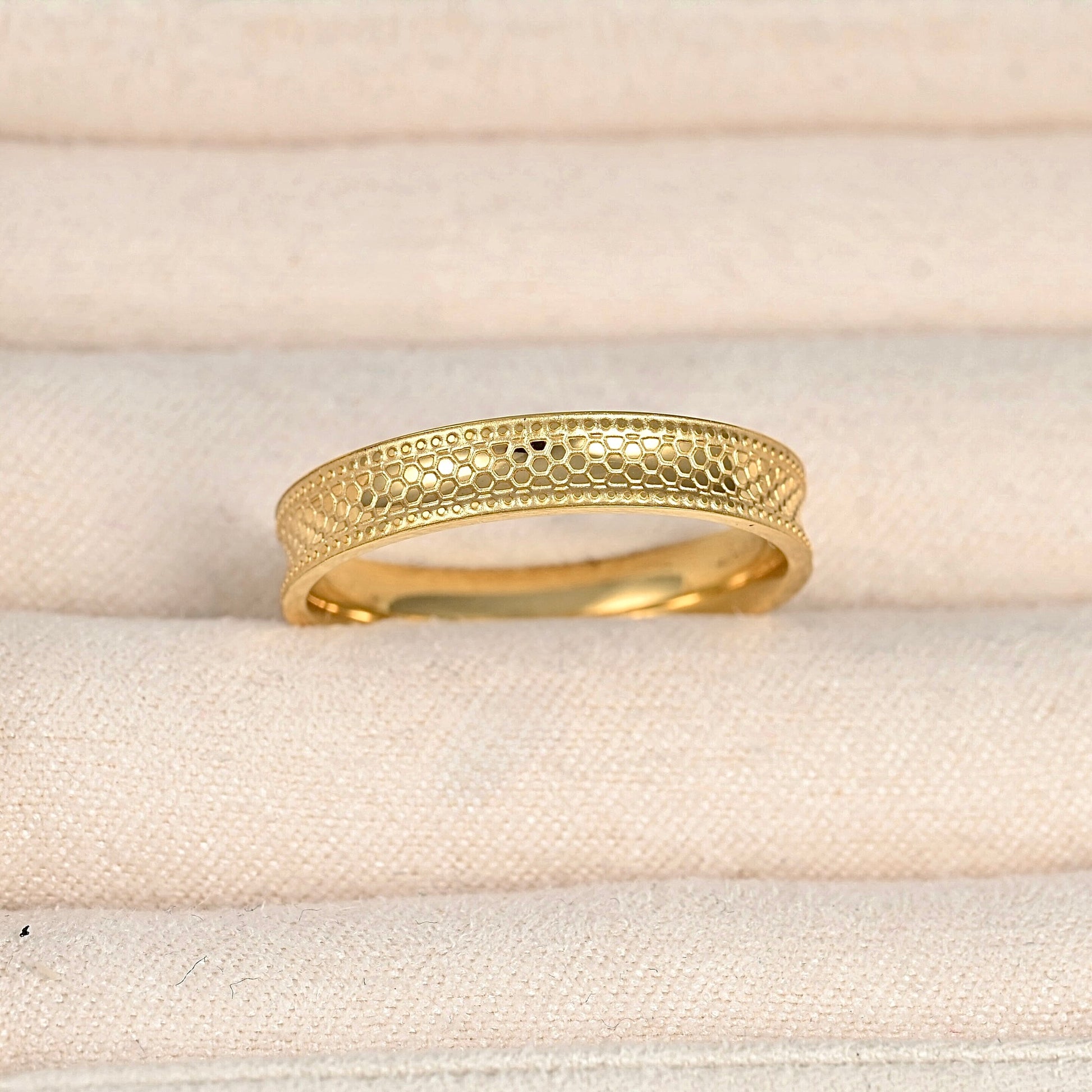 A gold ring with a textured honeycomb pattern on the top band, displayed on a beige fabric ring box.
