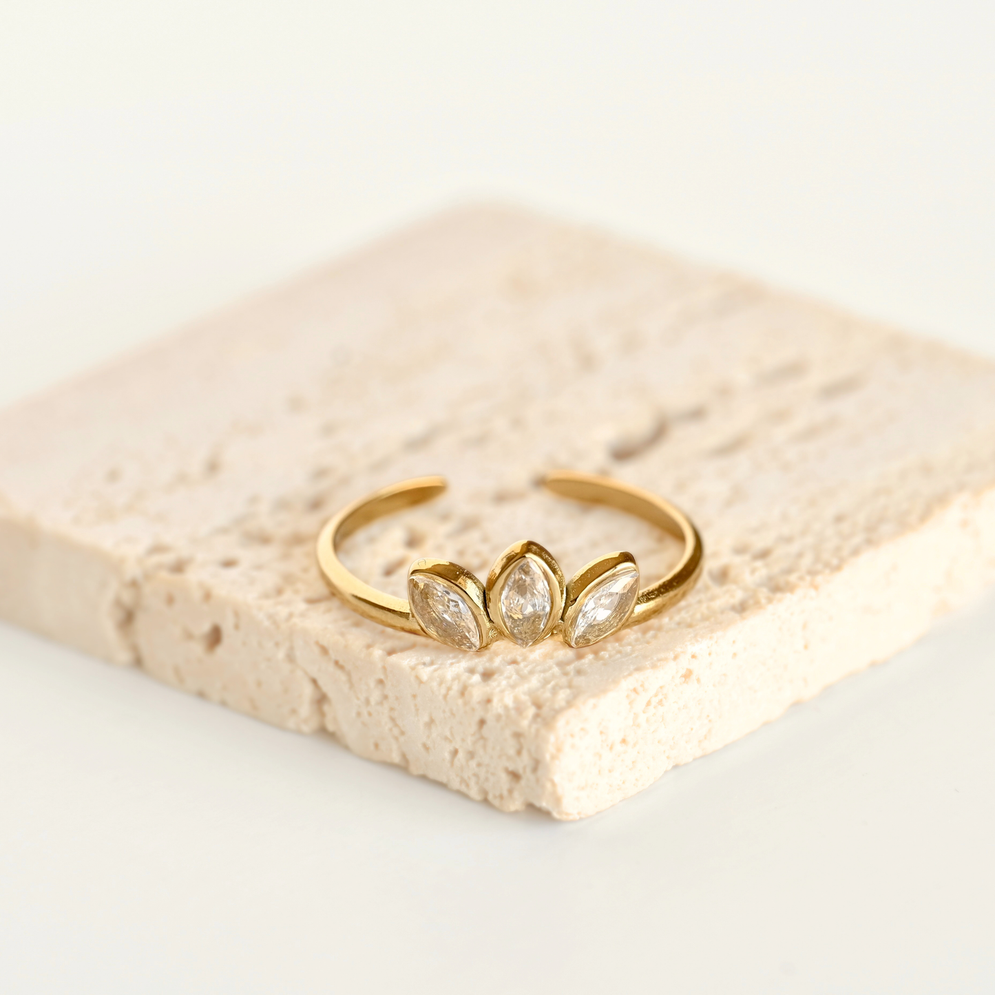 A gold open-ended ring with three marquise-cut clear gemstones, displayed on a textured beige stone tile against a white background.