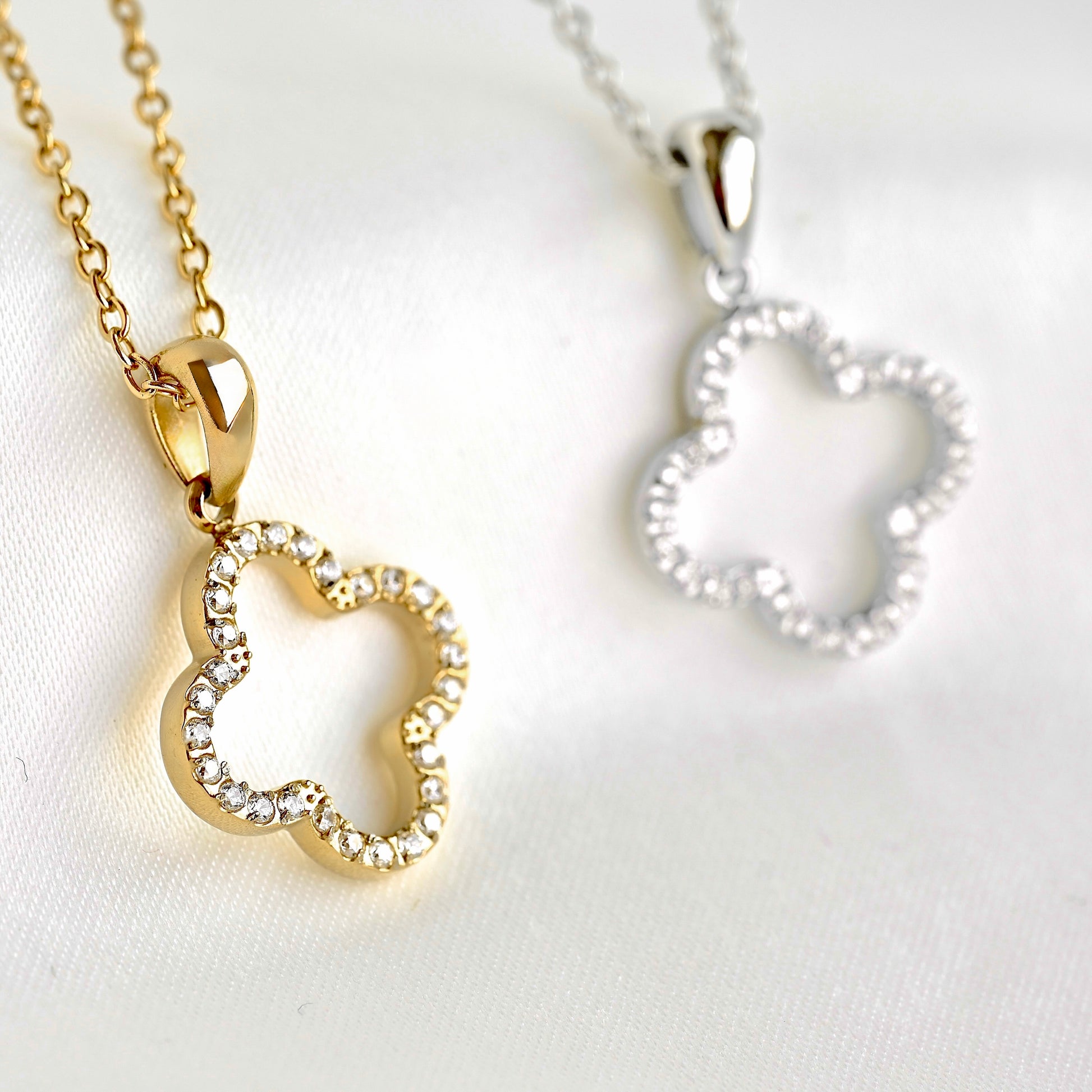 Two clover-shaped pendant necklaces on chains, one gold-tone with clear rhinestones and one silver-tone with clear rhinestones, displayed on a white fabric background.