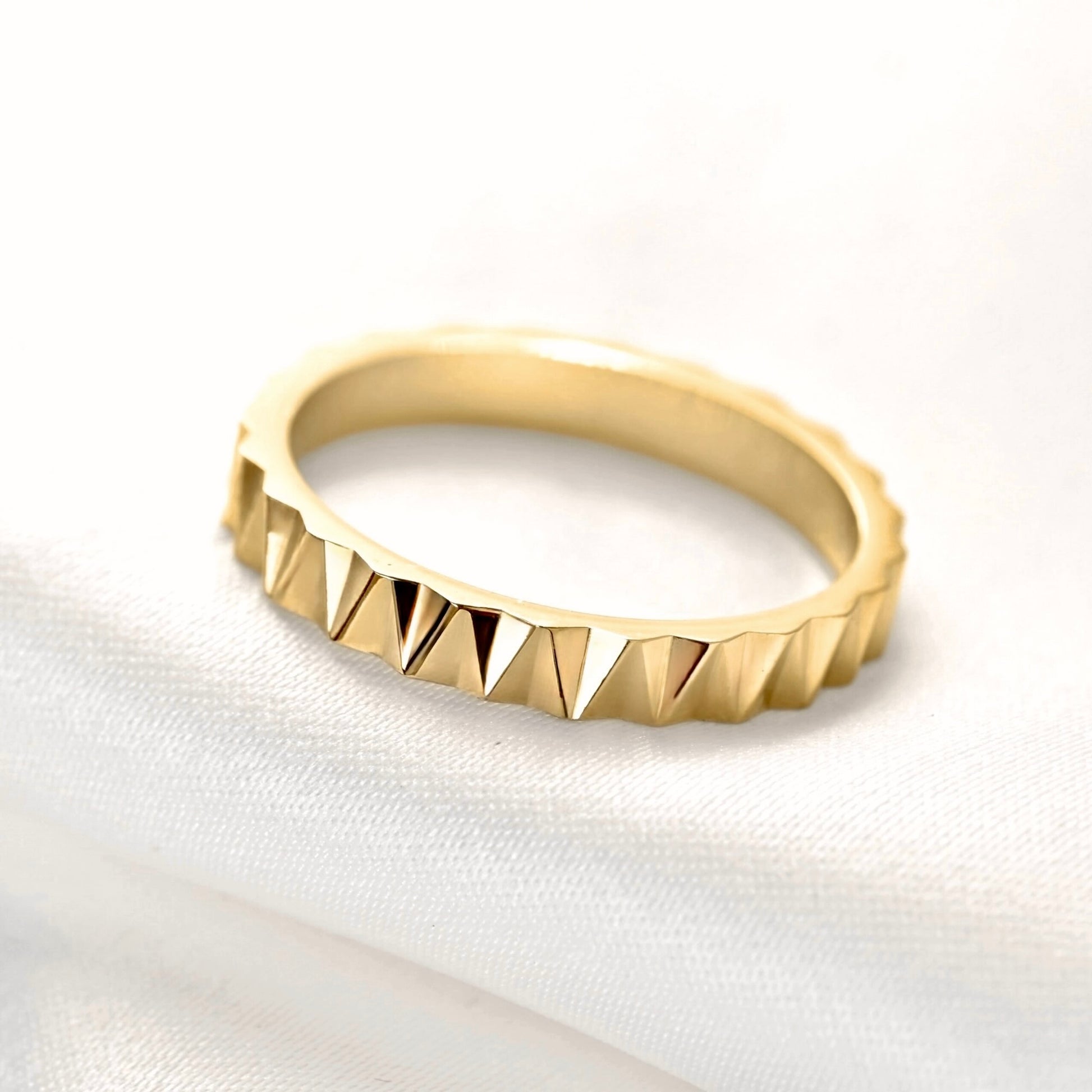 A gold ring with a textured, faceted triangular pattern around the outer edge, resting on a soft white fabric background.