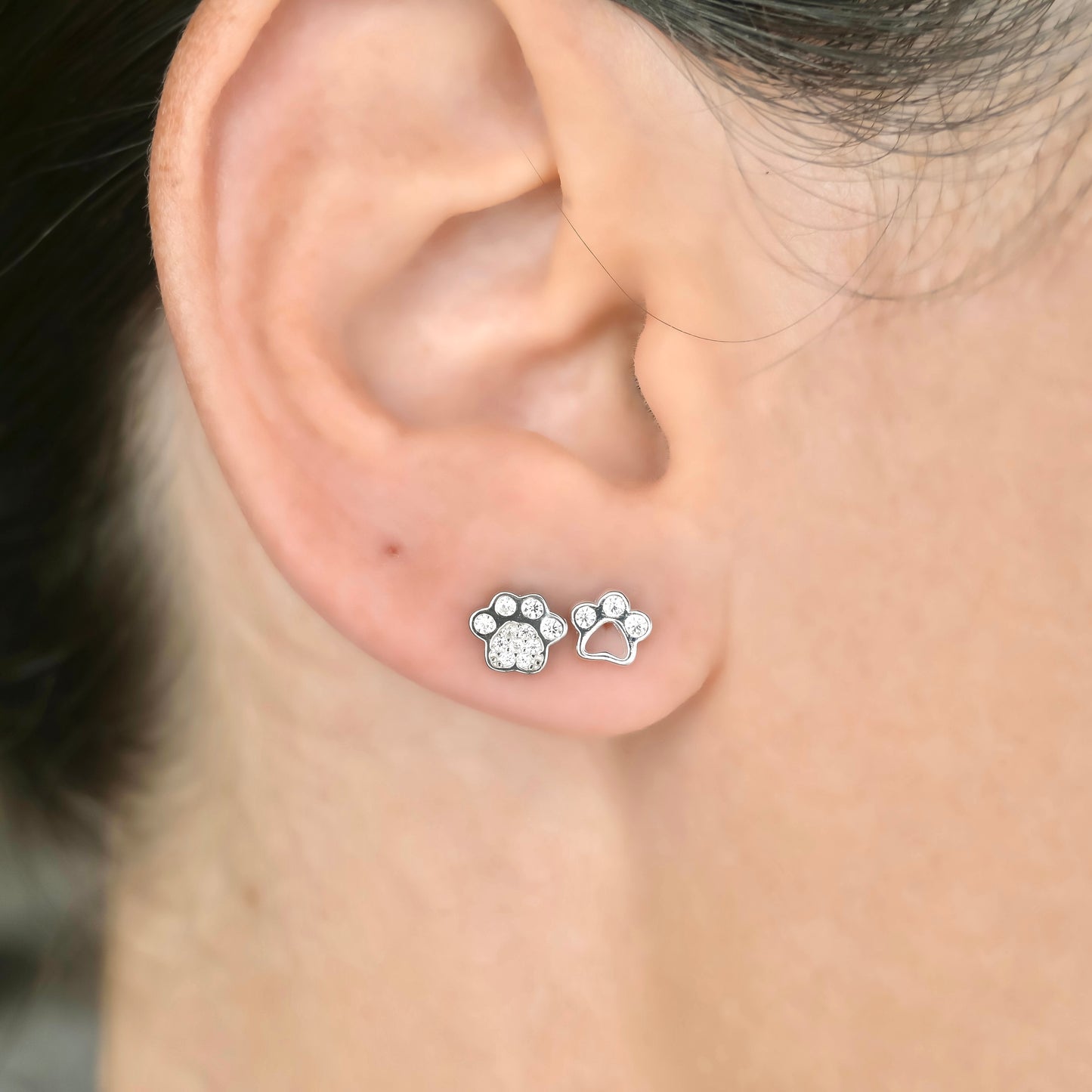 Close-up of an ear wearing two silver paw print stud earrings, one solid with pave CZ, the other cutout with three CZ stones