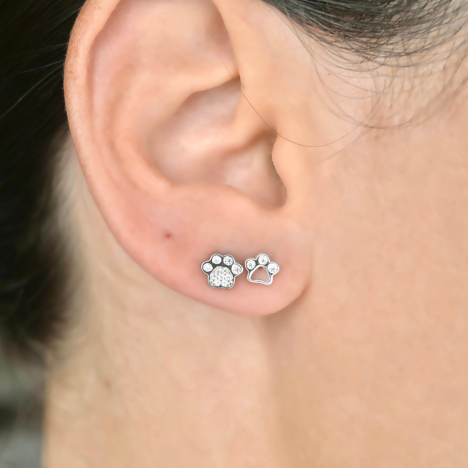 Close-up of an ear wearing two silver paw print stud earrings, one solid with pave CZ, the other cutout with three CZ stones