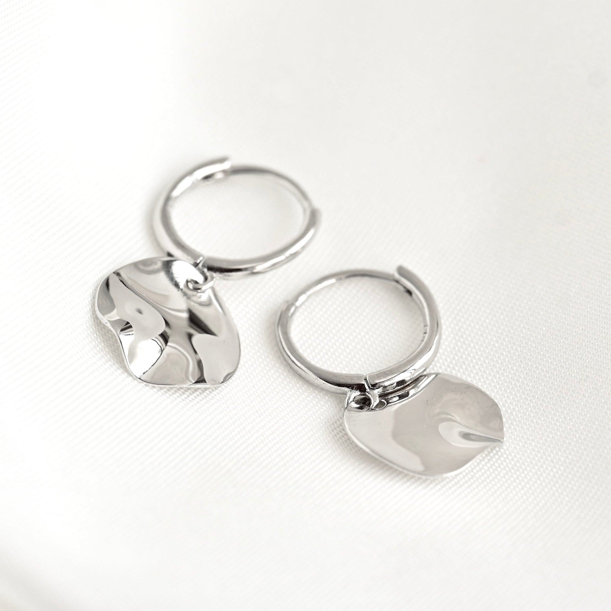 A pair of polished silver hoop earrings with irregular, organic-shaped dangling charms, displayed on a clean white textured fabric background.
