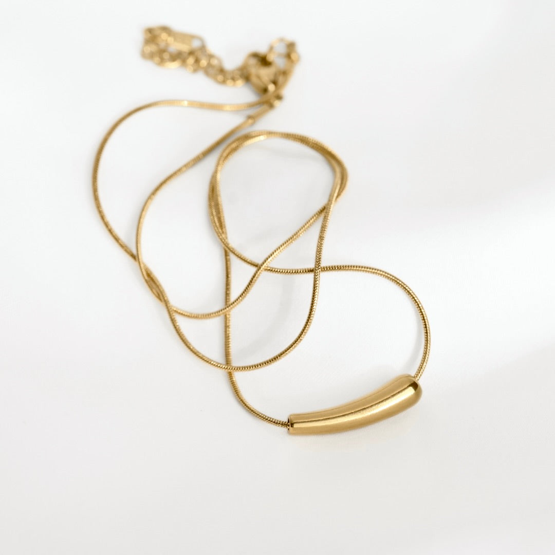A gold-toned snake chain necklace with a polished curved cylindrical pendant, displayed on a clean white background.