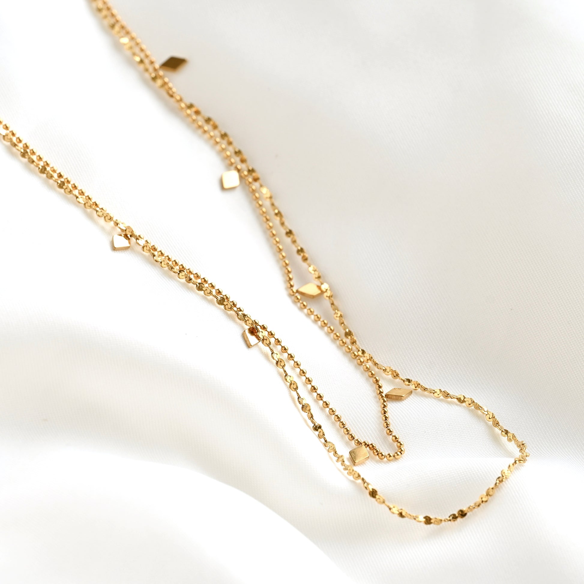 A delicate gold double-layer necklace with small diamond-shaped charms, laid out on a soft white fabric background.