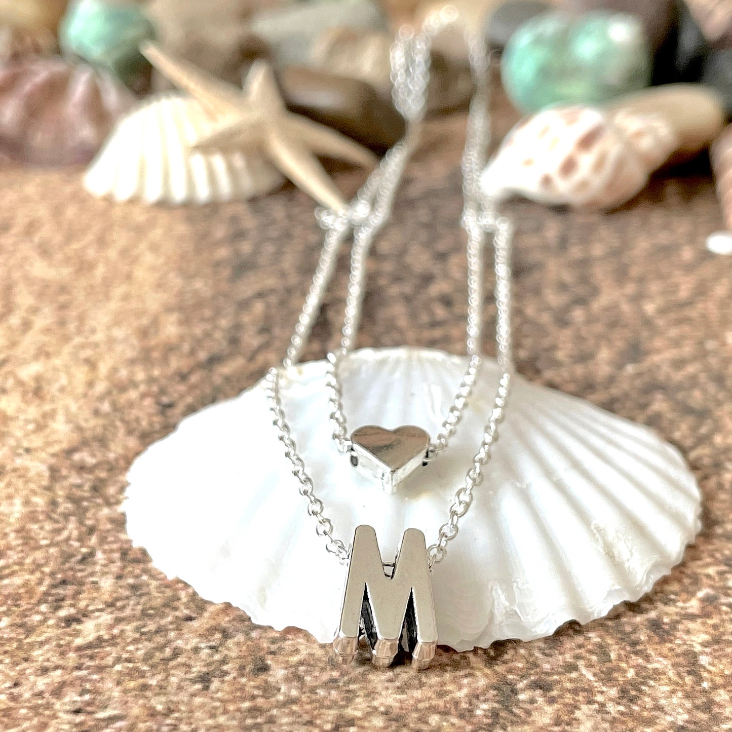 Double layered silver necklace with an 'M' and heart pendant on a seashell with starfish and shells in the background.