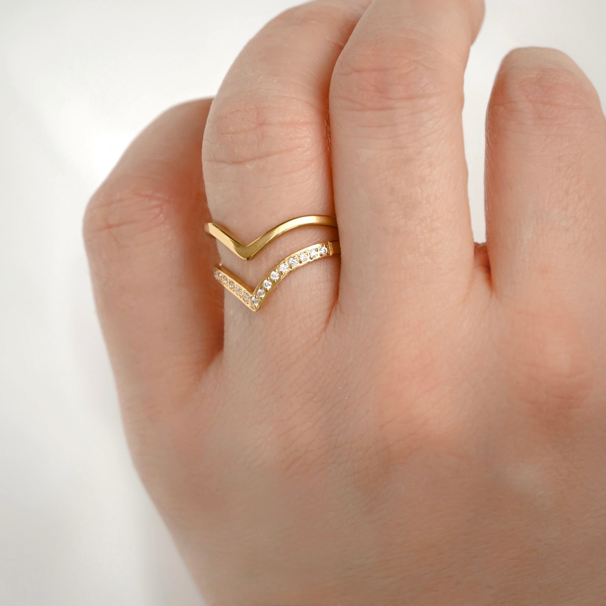 A close-up of a hand wearing a gold chevron ring with one flat gold chevron and one pavé-set chevron, photographed against a soft light gray background.