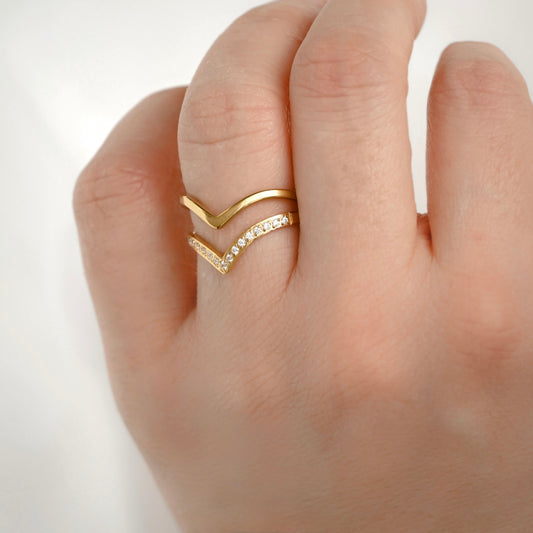 A close-up of a hand wearing a gold chevron ring with one flat gold chevron and one pavé-set chevron, photographed against a soft light gray background.