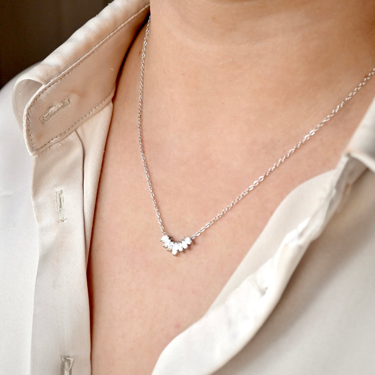 A close-up of a delicate silver necklace with a small curved cluster of clear stones on a chain, worn by a model in a white satin shirt.