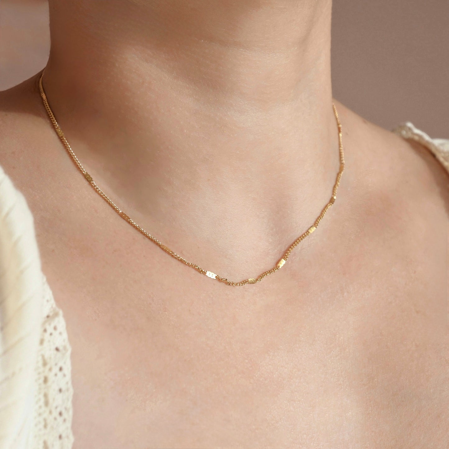 A delicate gold chain necklace with small flat rectangular links, worn on a model's neck over a cream-colored lace top.