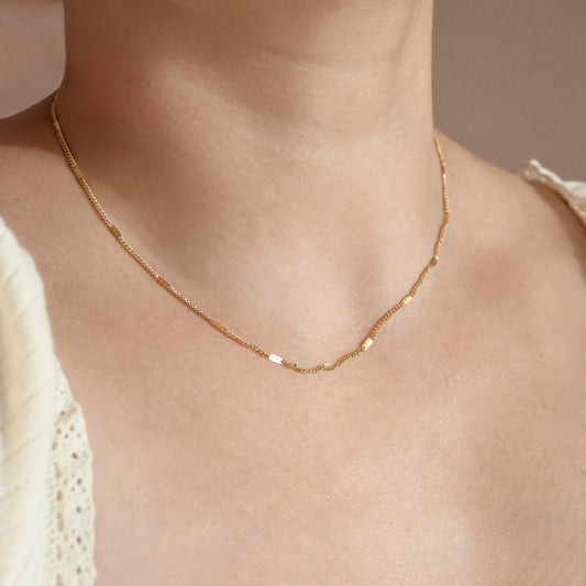 A delicate gold chain necklace with small flat rectangular links, worn on a model's neck over a cream-colored lace top.