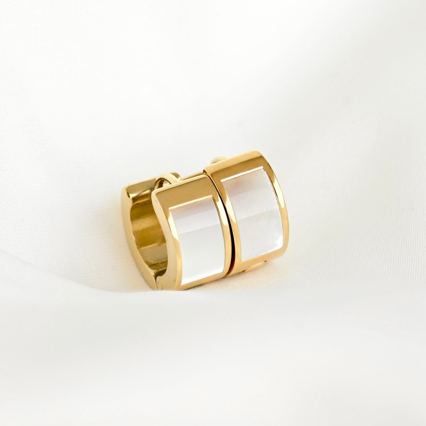 A gold-toned thick huggie earrings featuring two smooth, rounded white inlay panels, photographed on a clean white fabric background.