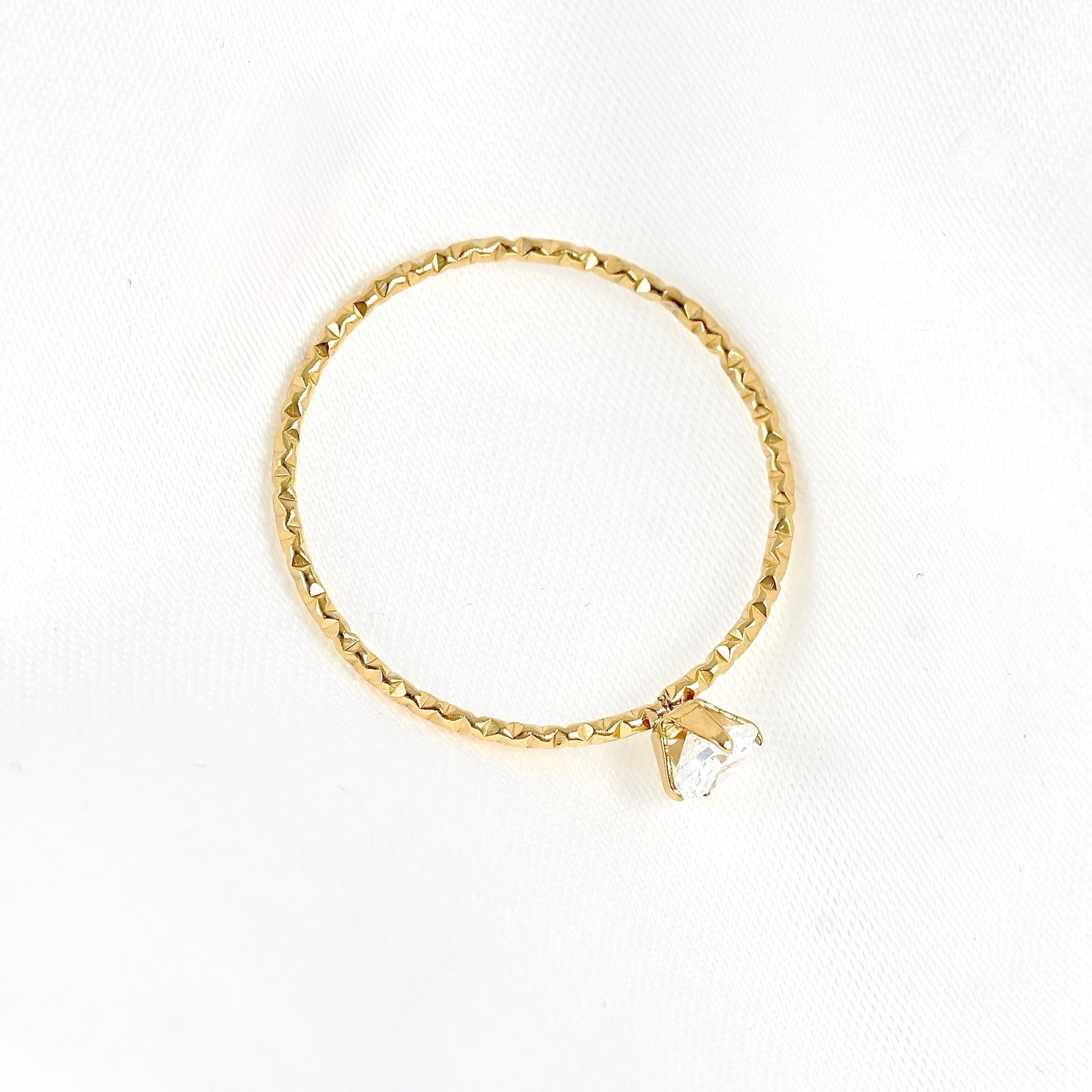 A delicate textured gold ring with a small clear stone, displayed on a textured white fabric background.