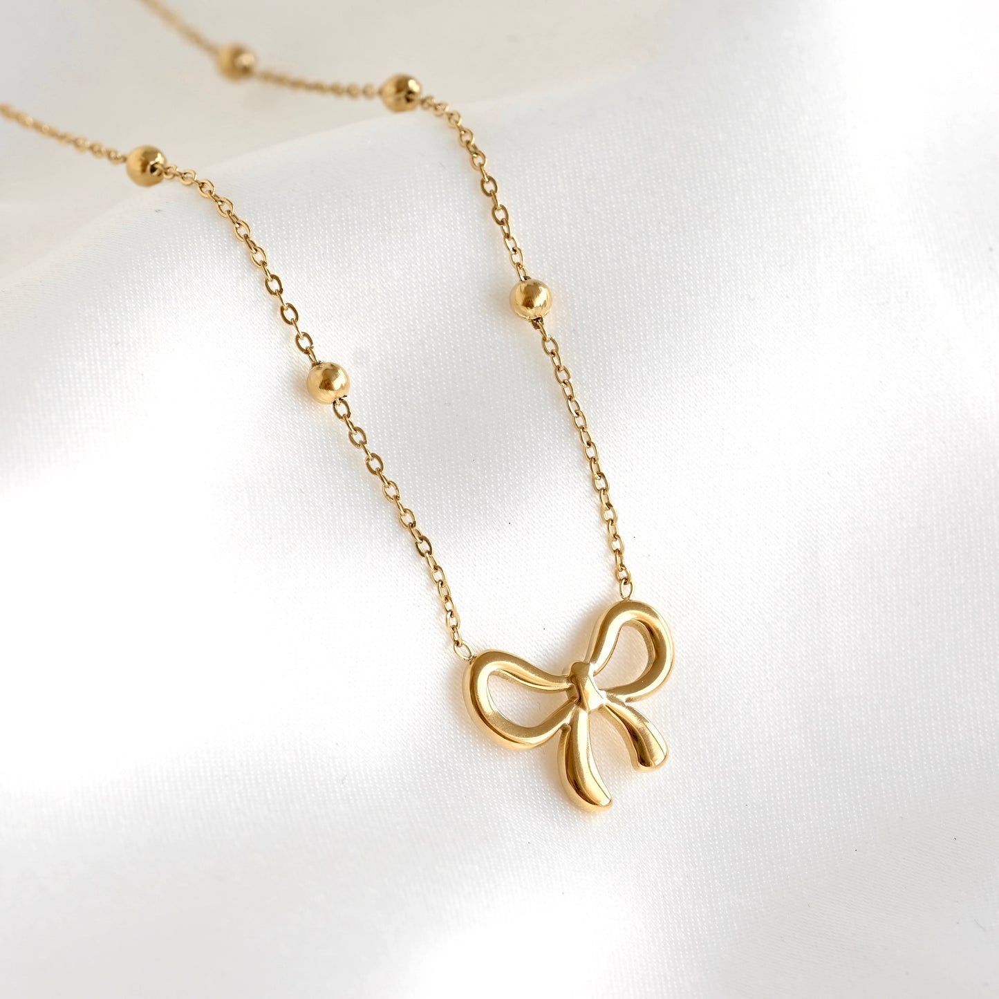 A delicate gold necklace featuring a small bow pendant and small bead accents on the chain, displayed on a soft white fabric background.