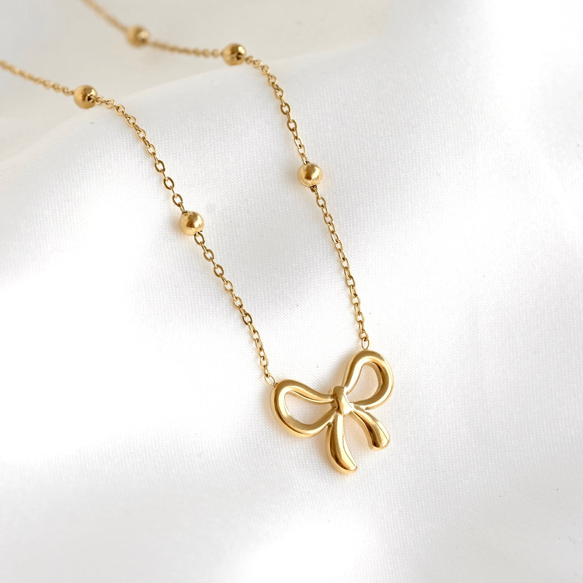 A delicate gold necklace featuring a small bow pendant and small bead accents on the chain, displayed on a soft white fabric background.