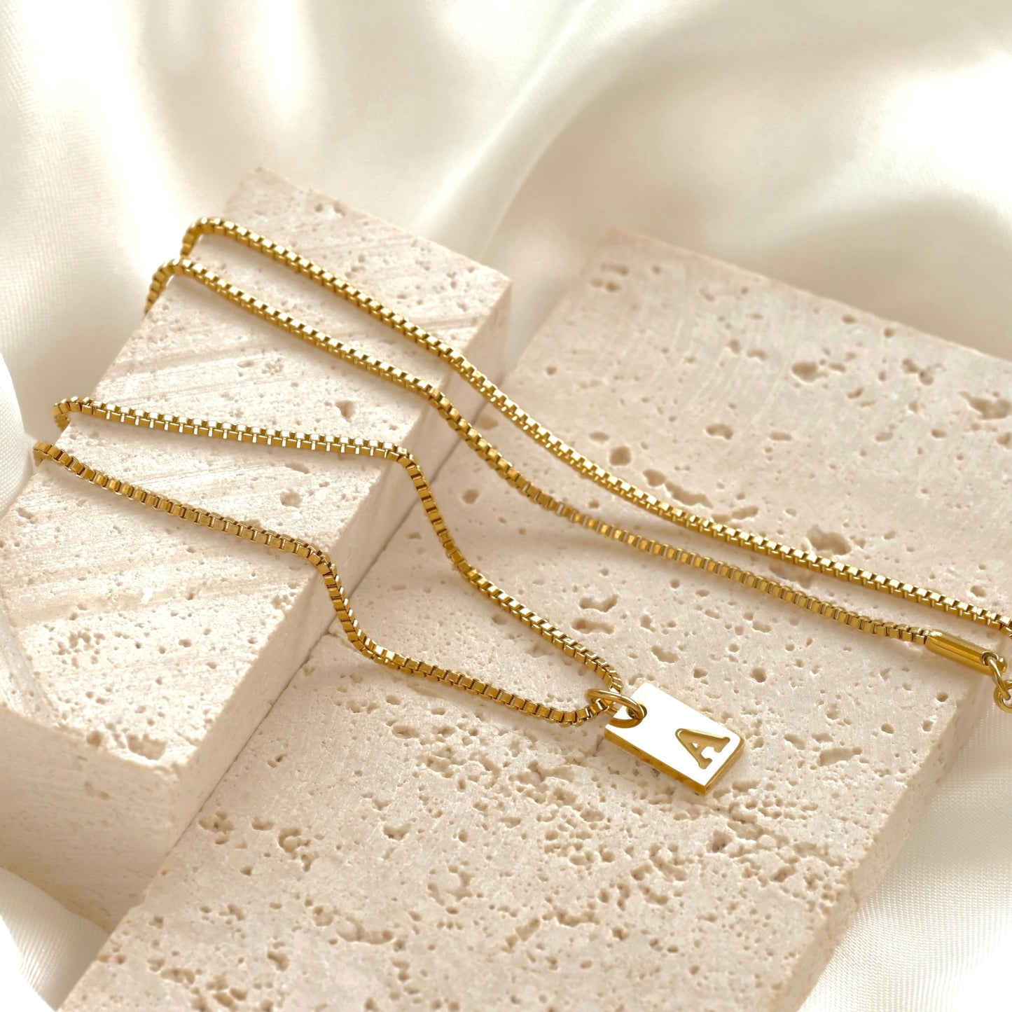 Gold box necklace with a rectangular initial charm on a textured stone surface