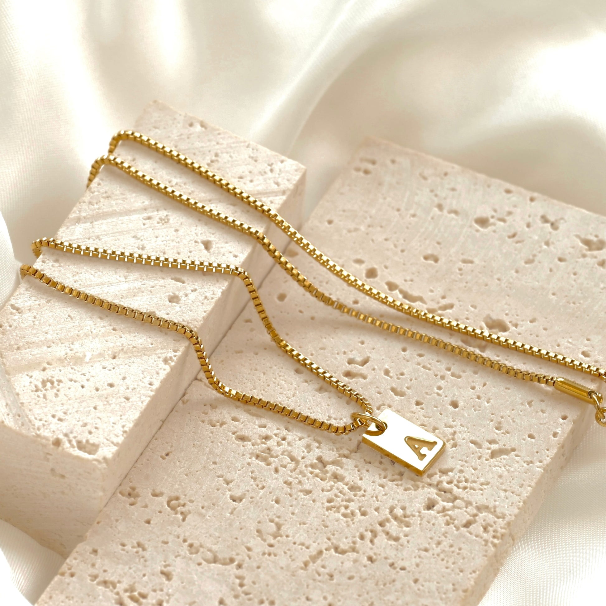 Gold box necklace with a rectangular initial charm on a textured stone surface