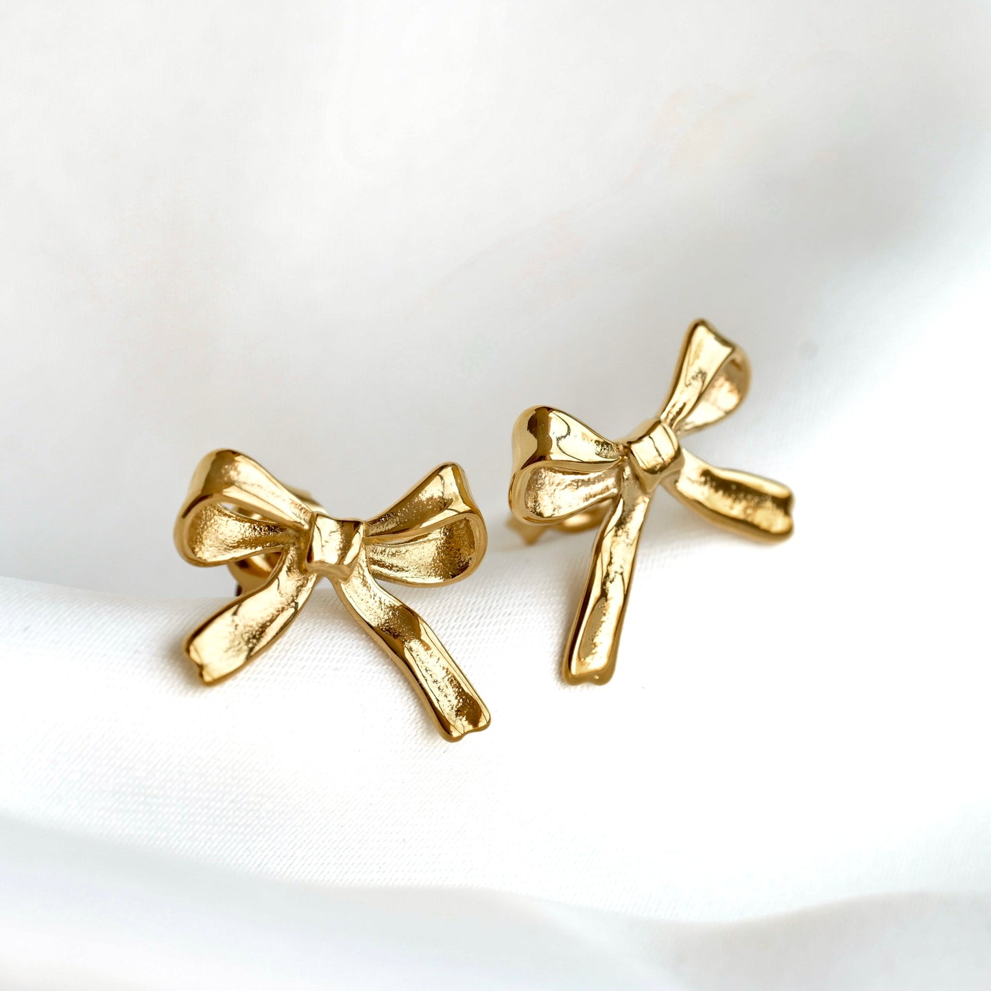A pair of gold bow-shaped stud earrings displayed on a soft, white draped fabric background.