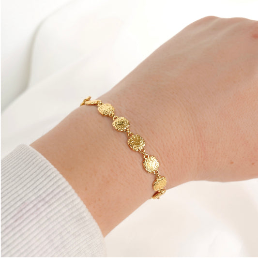 A delicate gold chain bracelet with textured, hammered square links worn on a person's wrist, shown against a soft white background.