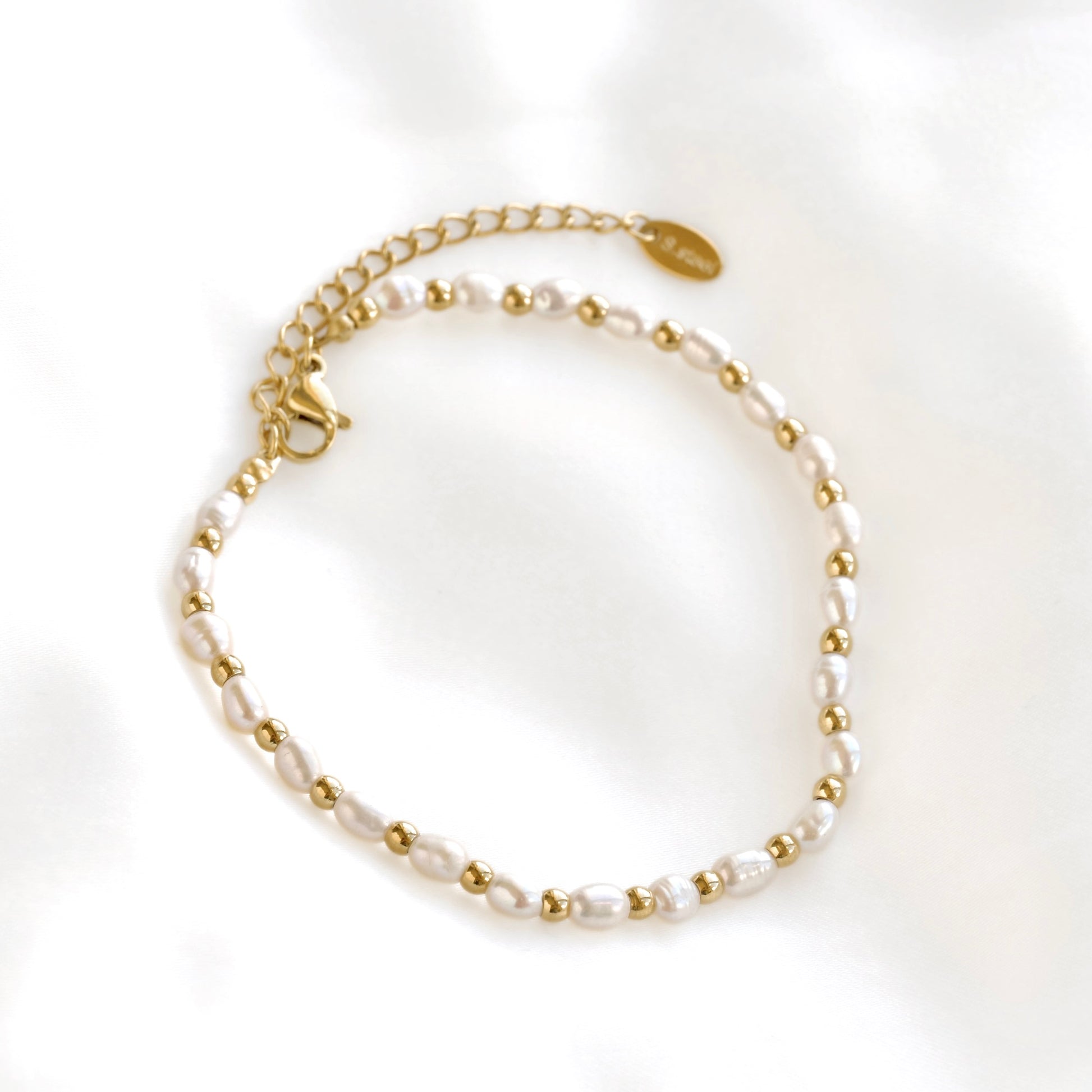 A delicate beaded bracelet featuring white baroque pearls and small gold spacer beads, connected by a gold chain extender with a lobster clasp and a small oval brand tag, displayed on a soft white fabric background.
