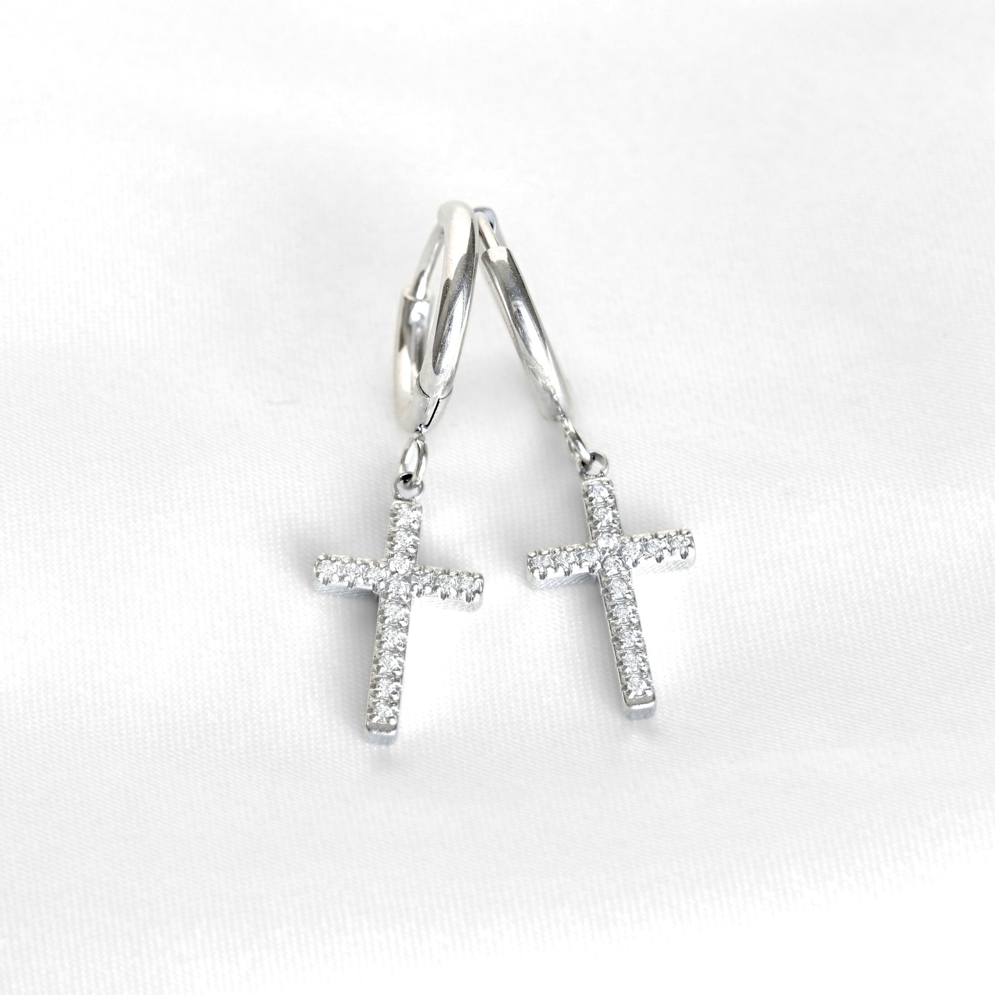 A pair of silver cross hoop earrings with small cross charms paved with clear stones, displayed on a soft white fabric background.