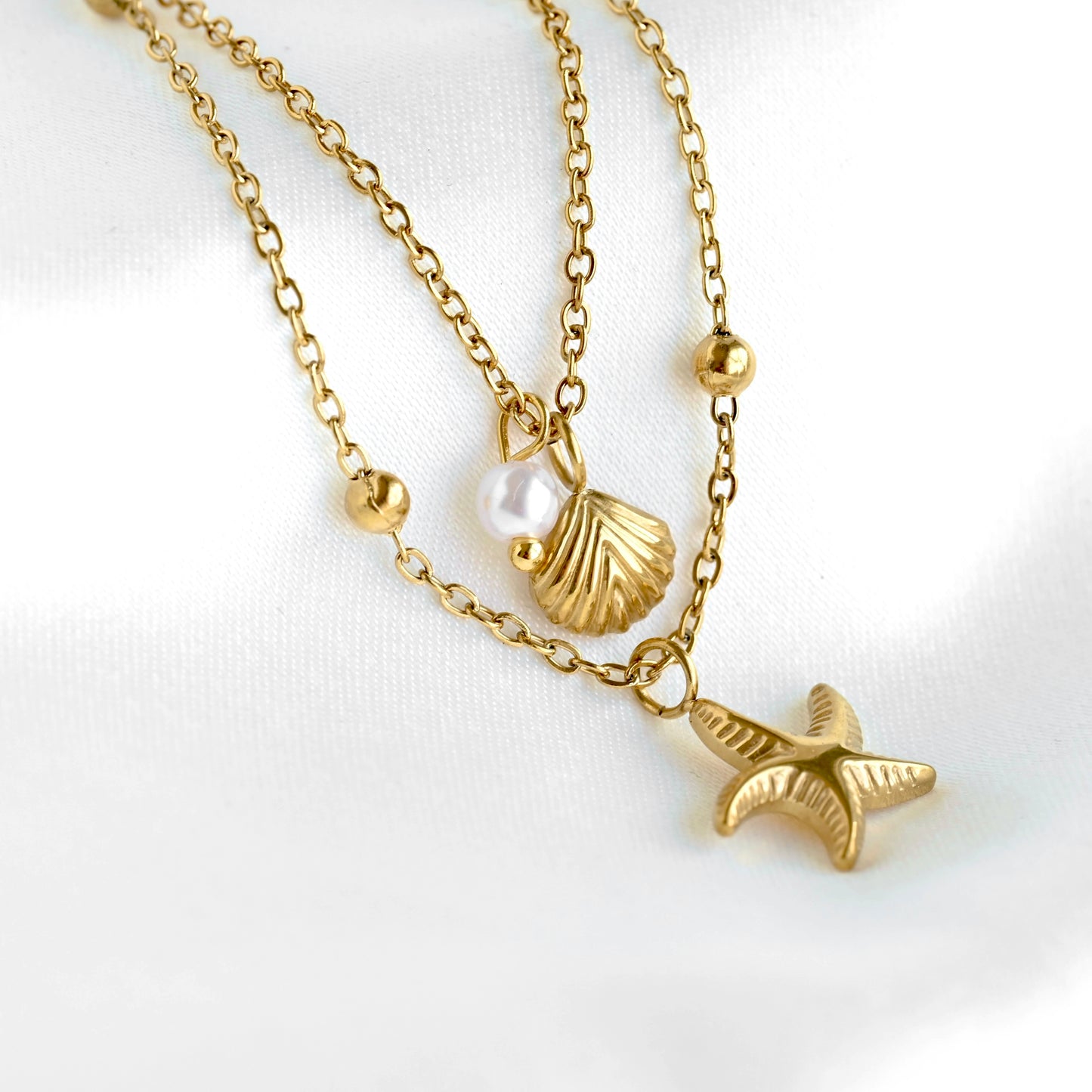 A gold-tone double-strand necklace featuring a starfish pendant, a textured seashell, and a small white pearl bead, displayed on a soft white fabric background.