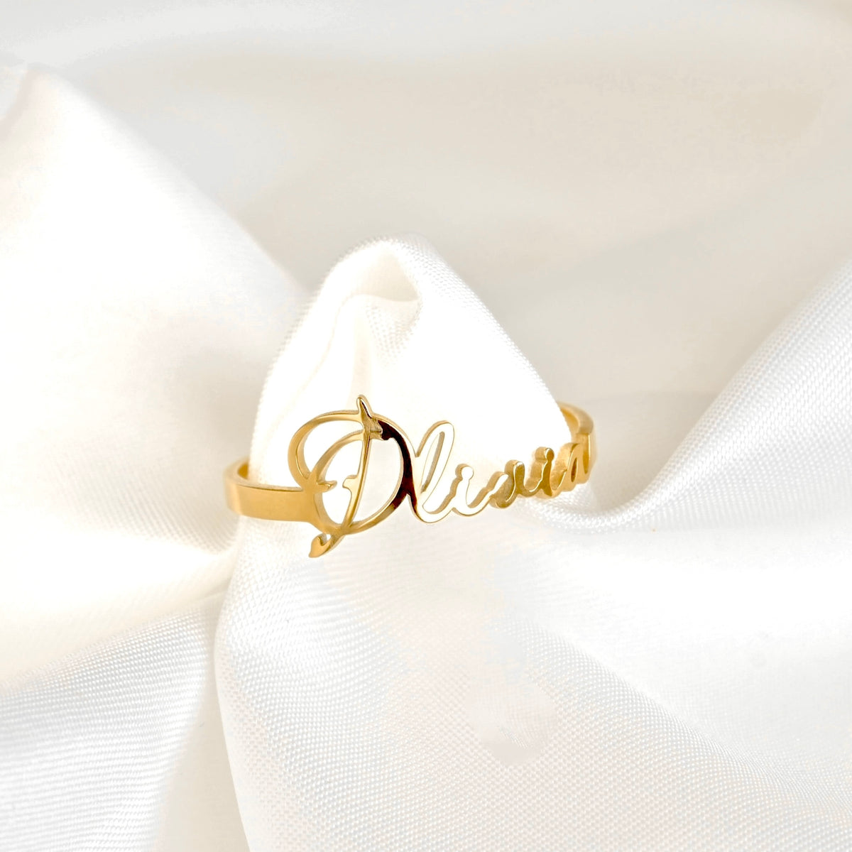 A gold script name ring reading 'Olivia' displayed on a white satin ribbon bow.