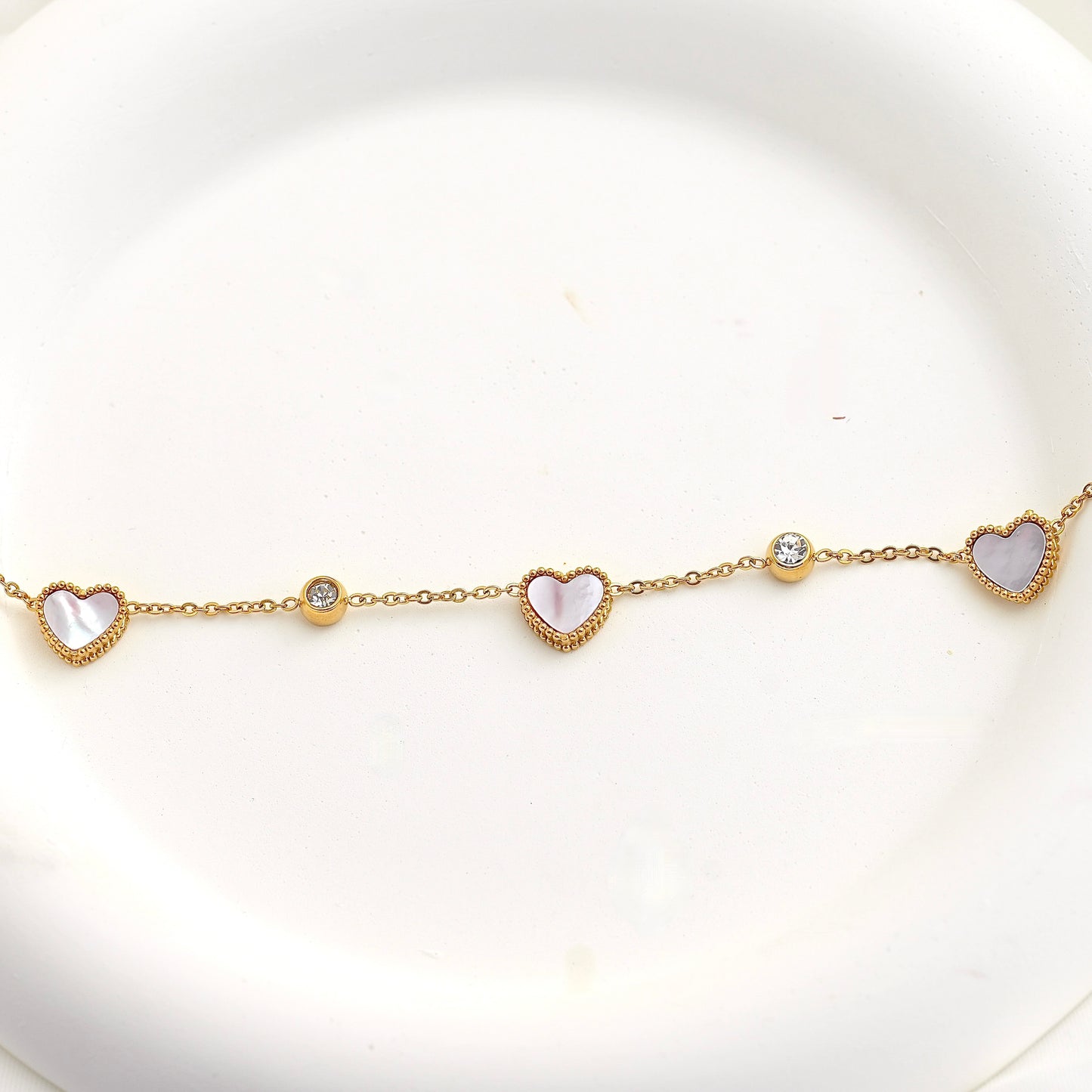 A delicate gold chain bracelet featuring three heart-shaped mother-of-pearl pendants and two small clear crystal beads, displayed on a white ceramic plate.
