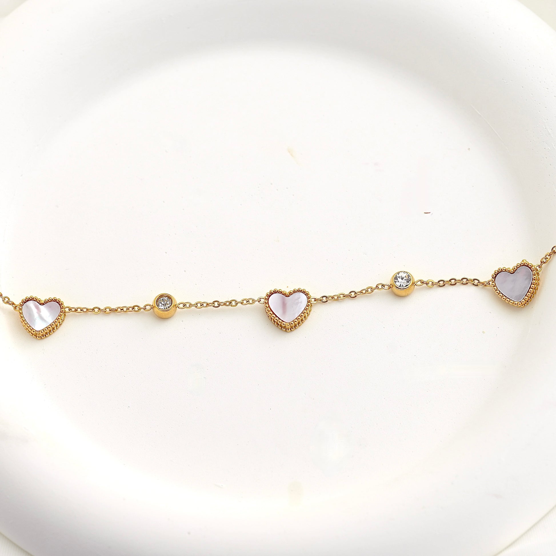 A delicate gold chain bracelet featuring three heart-shaped mother-of-pearl pendants and two small clear crystal beads, displayed on a white ceramic plate.