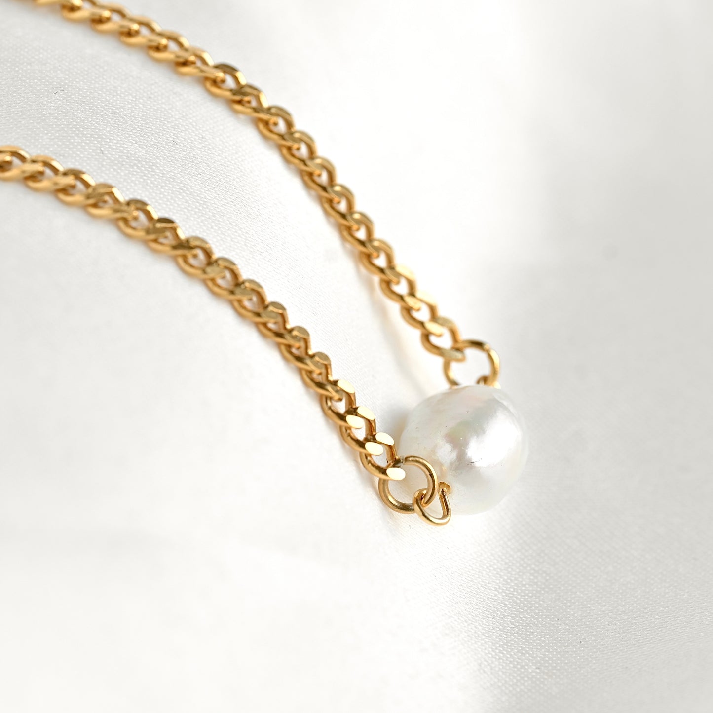 A close-up of a gold chain necklace with a single white pearl pendant resting on a white fabric background.