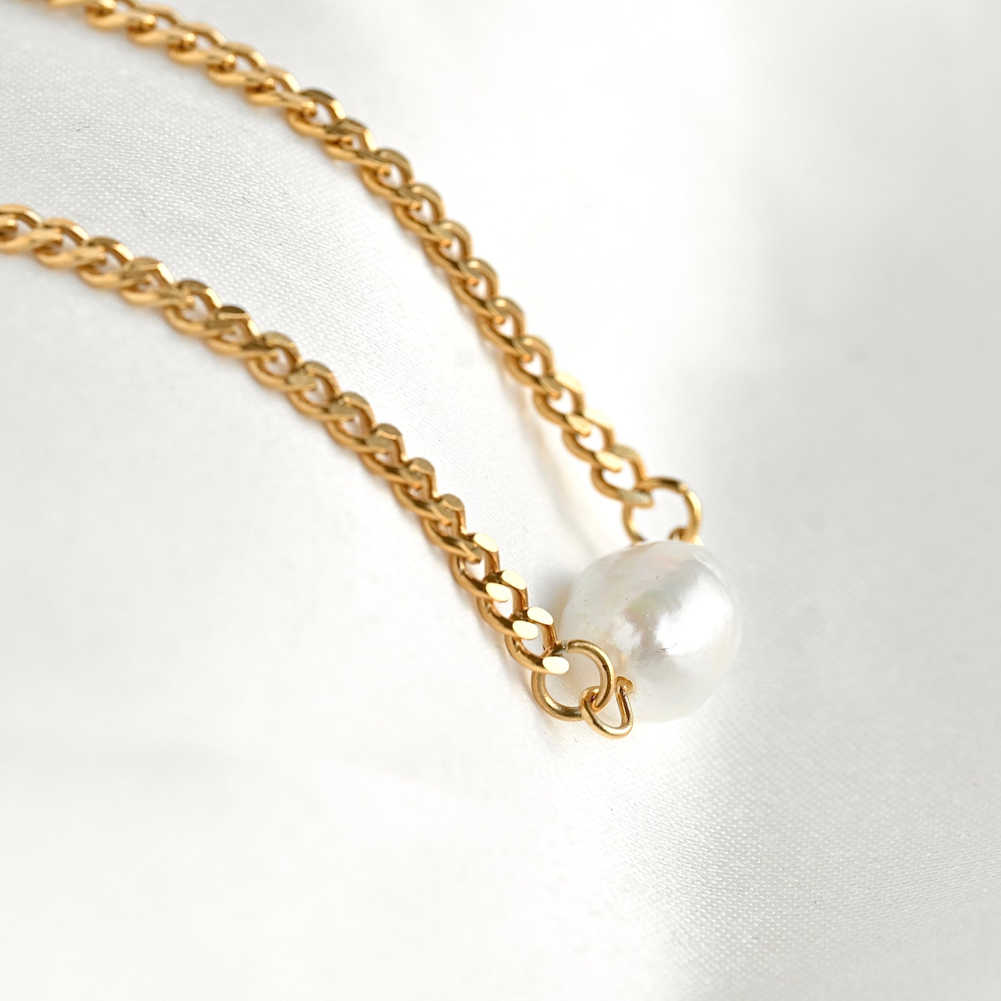 A close-up of a gold chain necklace with a single white pearl pendant resting on a white fabric background.