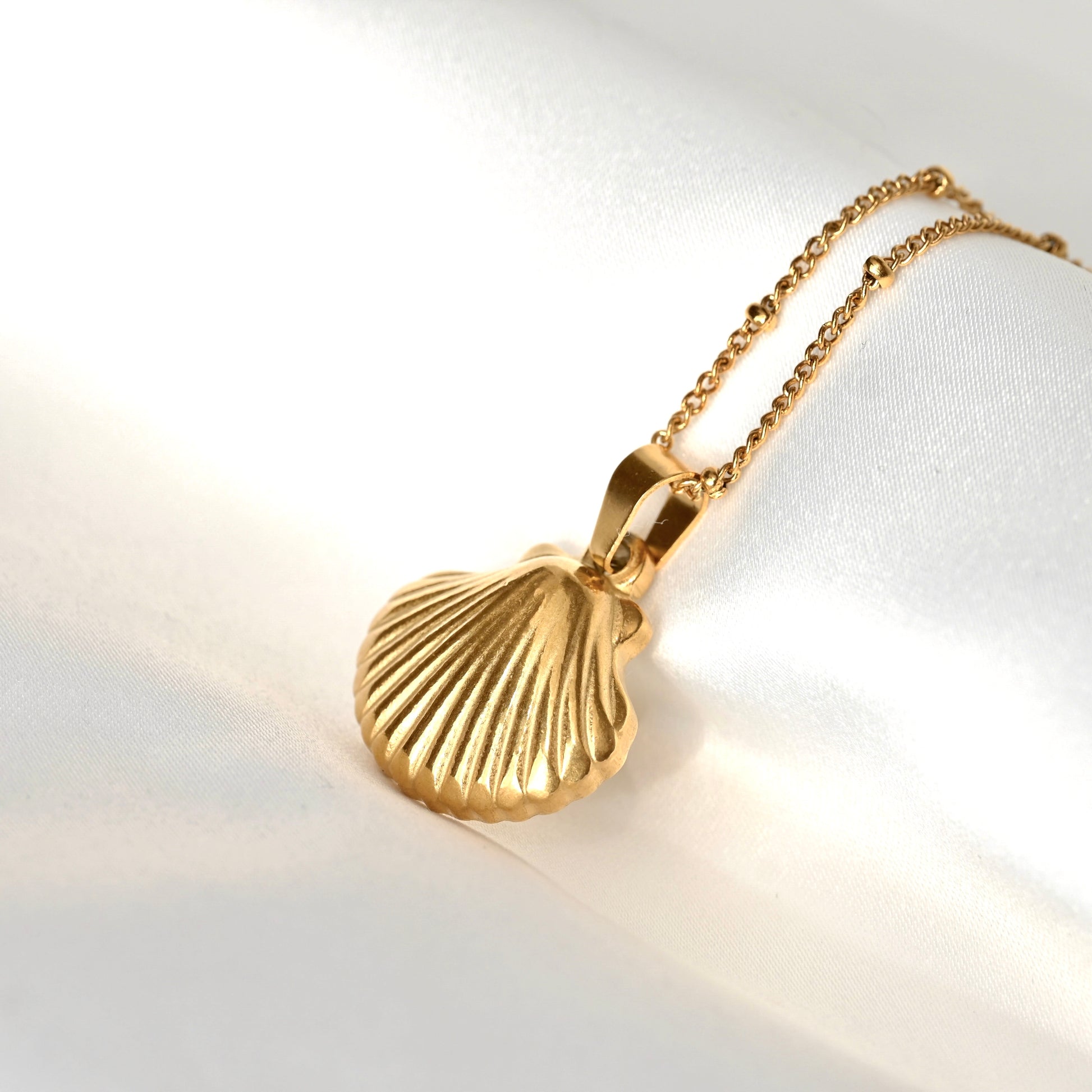 A gold shell-shaped pendant necklace on a delicate gold chain, displayed on a soft white satin fabric background.