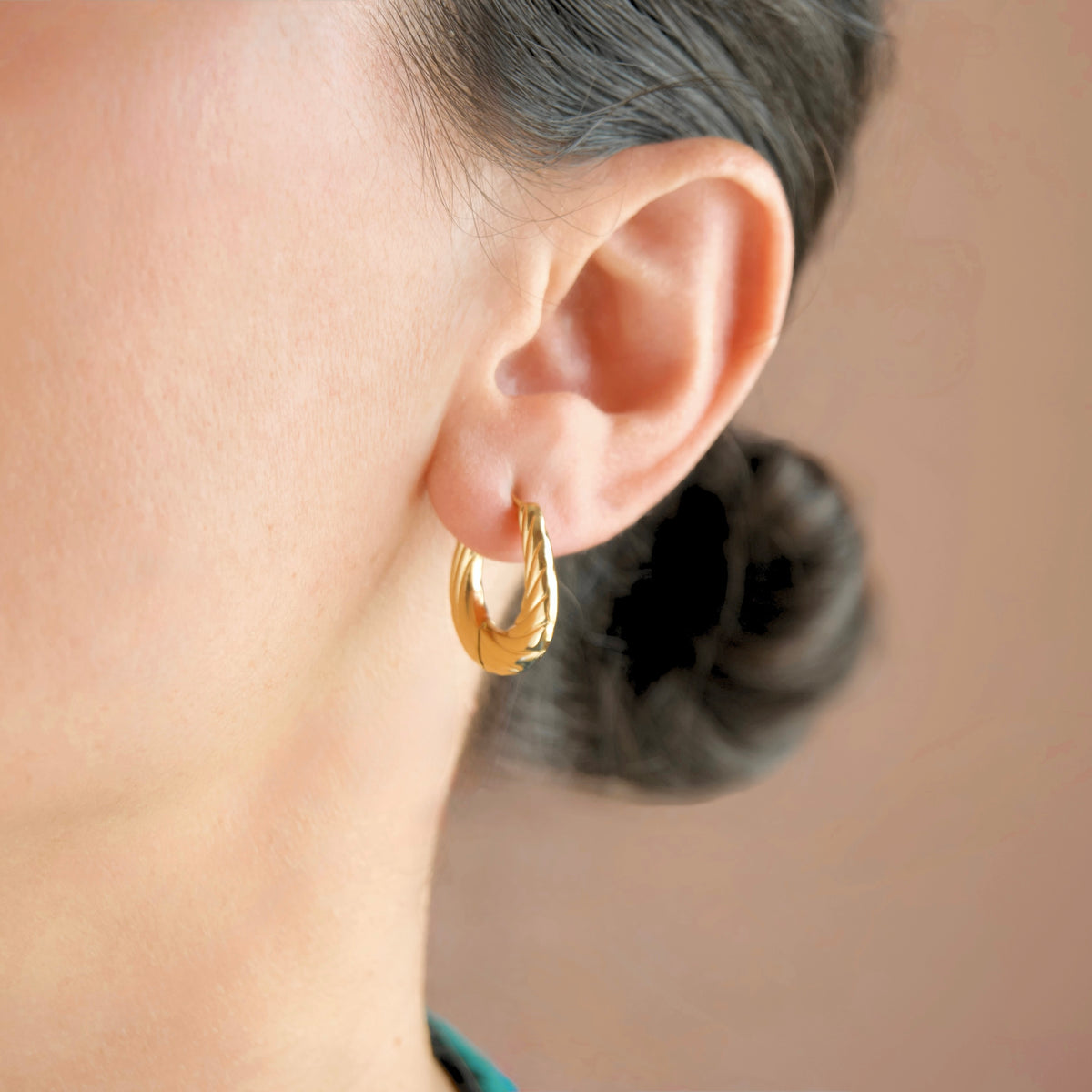 A close-up of a person's ear wearing a small twisted gold hoop earring against a soft beige background.