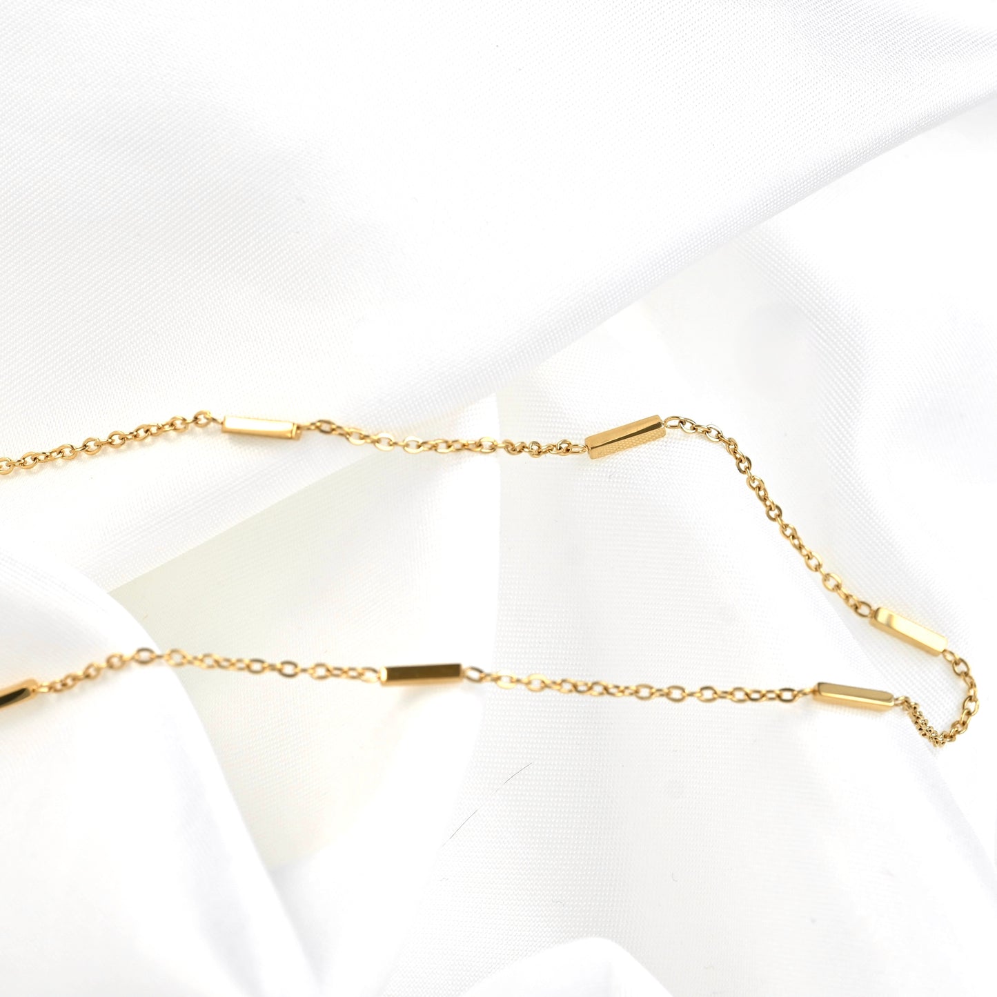A delicate gold chain necklace with small polished rectangular bar accents, laid out on a soft white fabric background.