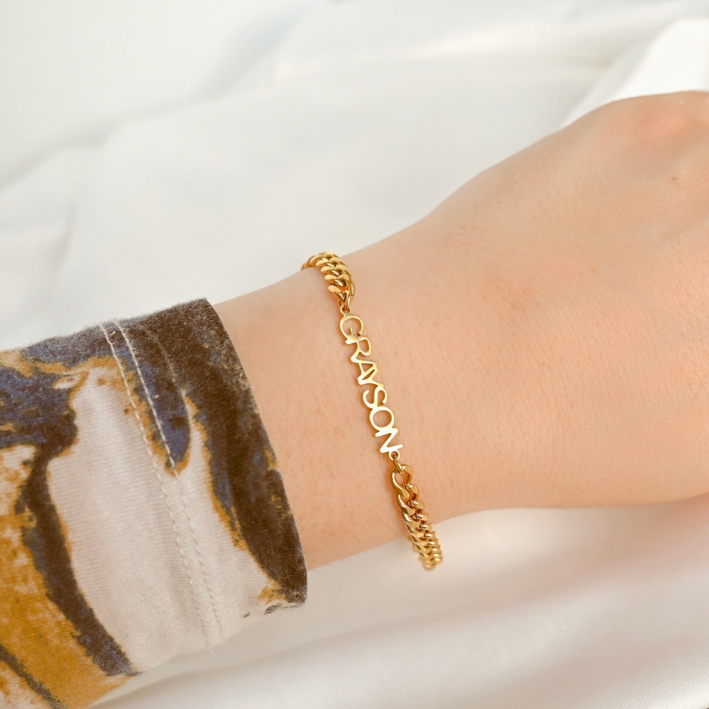 A gold curb chain bracelet with the name 'GRAYSON' spelled out in linked letters, worn on a person's wrist against a soft white fabric background.