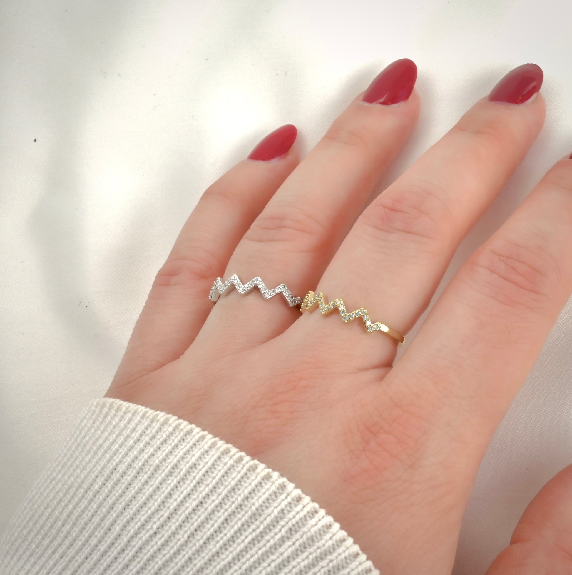 Hand wearing two zigzag pattern rings, one gold and one silver, side by side, on a plain background