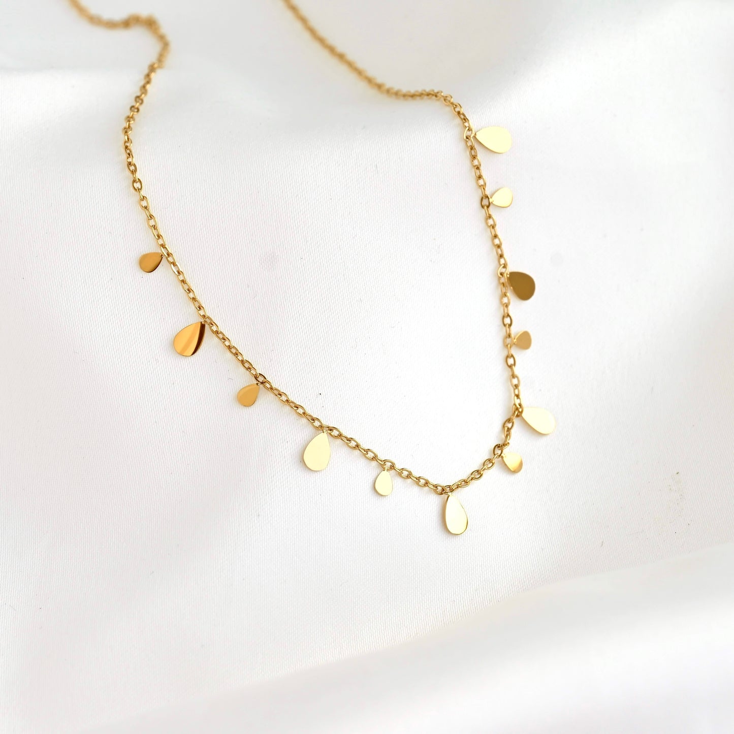 A delicate gold chain necklace with small, irregular flat disc and teardrop charms, displayed on a soft white fabric background.