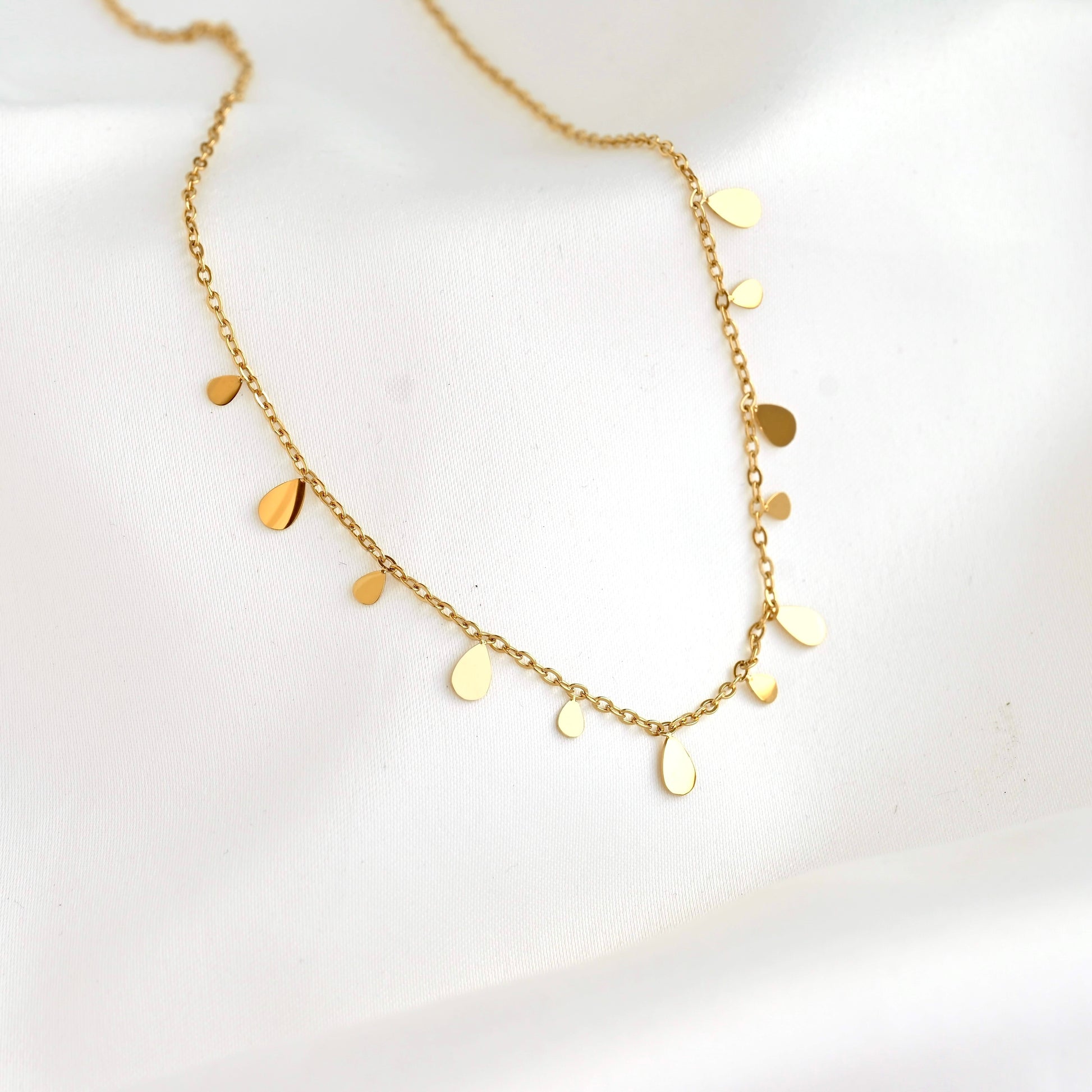 A delicate gold chain necklace with small, irregular flat disc and teardrop charms, displayed on a soft white fabric background.