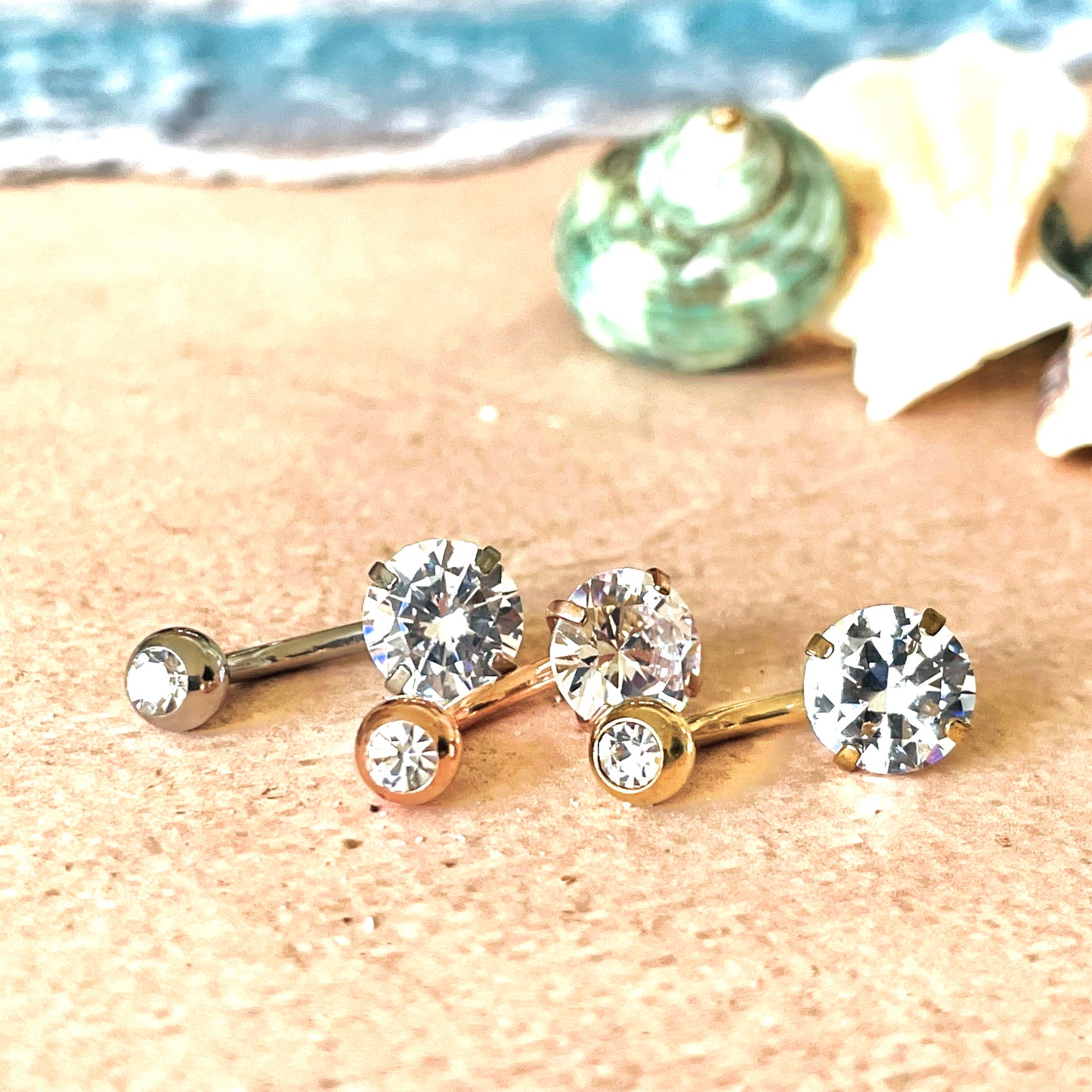 Gold diamond belly button rings in gold, silver and rose gold with clear gemstones on a sandy surface with shells in the background