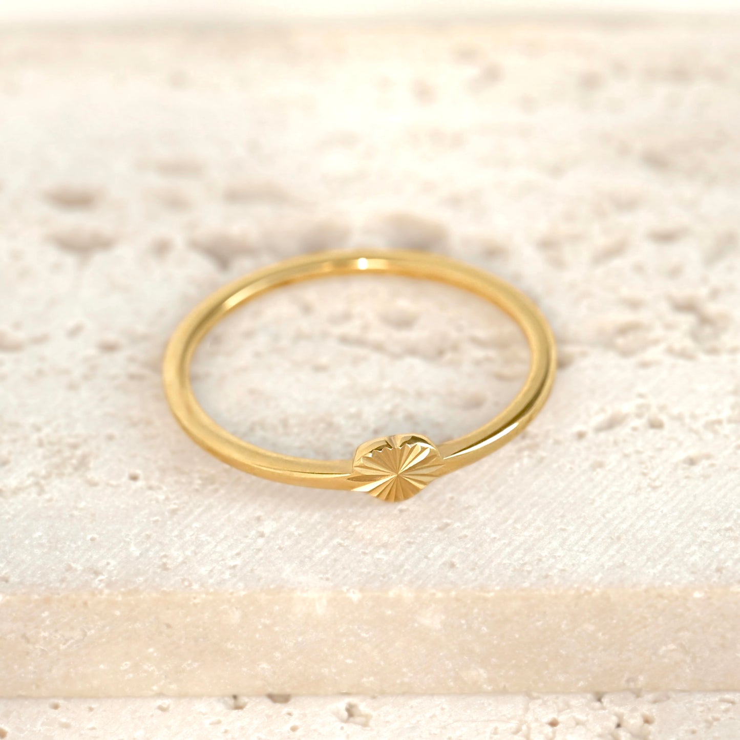 A delicate gold ring featuring a small sunburst heart detail on the front, resting on a textured, light beige stone surface.